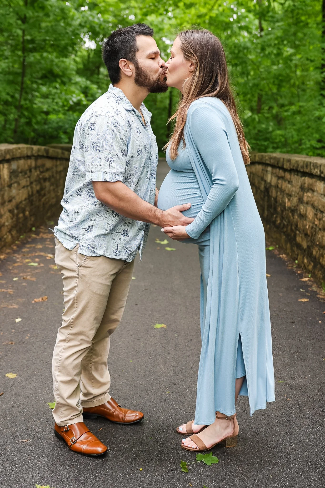 A man and woman are standing face-to-face outdoors, kissing, with the man holding the woman’s belly. The groom is dressed in beige pants, a patterned short-sleeved shirt, and brown shoes. The woman is wearing a long, light blue dress and tan high-hee