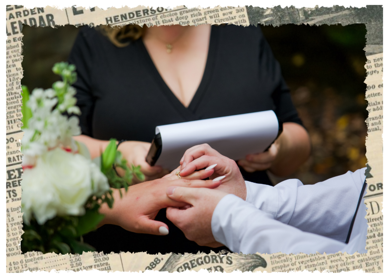 A couple holding hands during a wedding ceremony, with the bride wearing a white dress and the groom in a white shirt. An officiant, dressed in black, is reading from a notepad, and there is a bouquet of white flowers visible in the foreground.