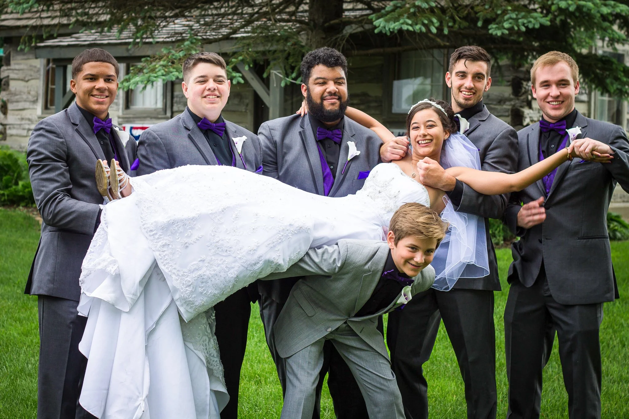 Group of six people, five men and one woman, in formal wedding attire outdoors, with a man in a tuxedo holding a woman in a wedding dress.