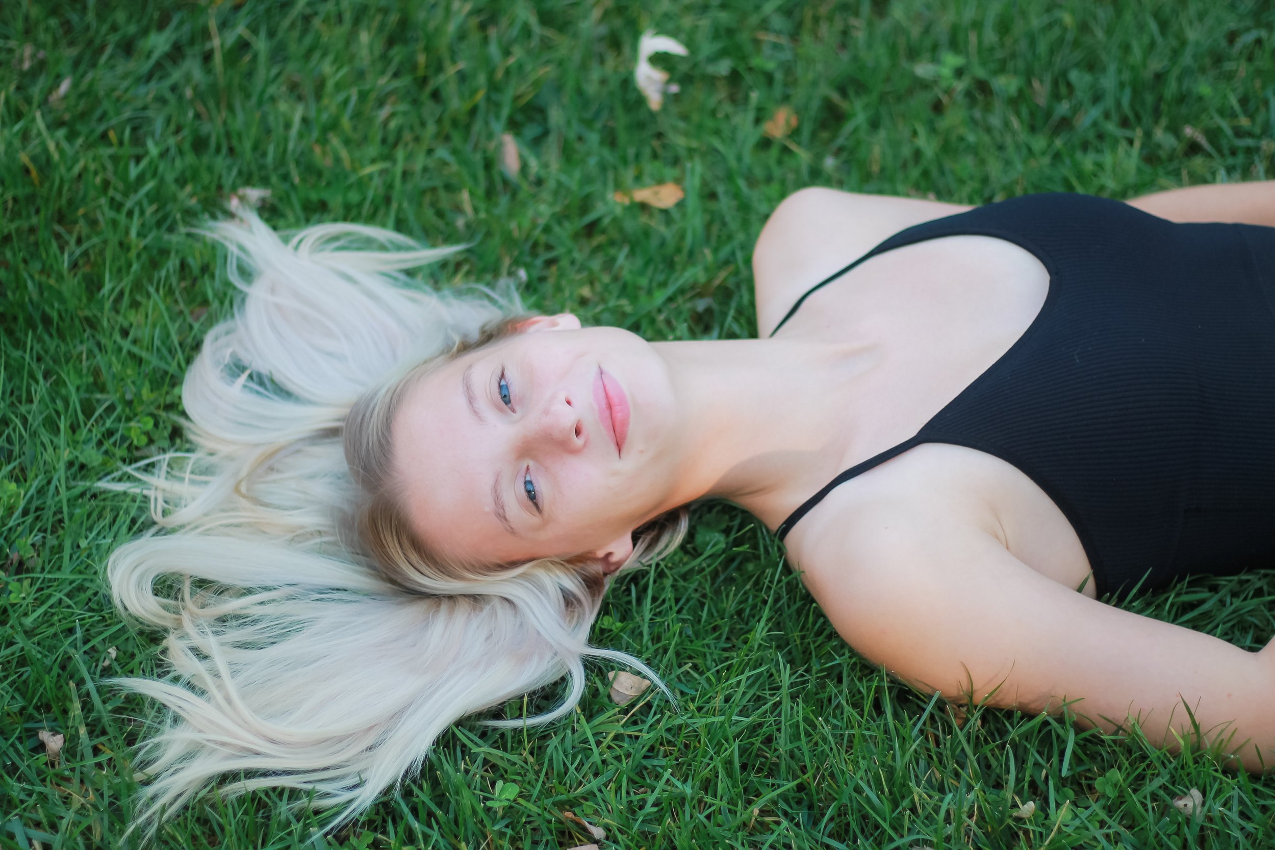 A young woman with long blonde hair and blue eyes lies on green grass, wearing a black sleeveless top.