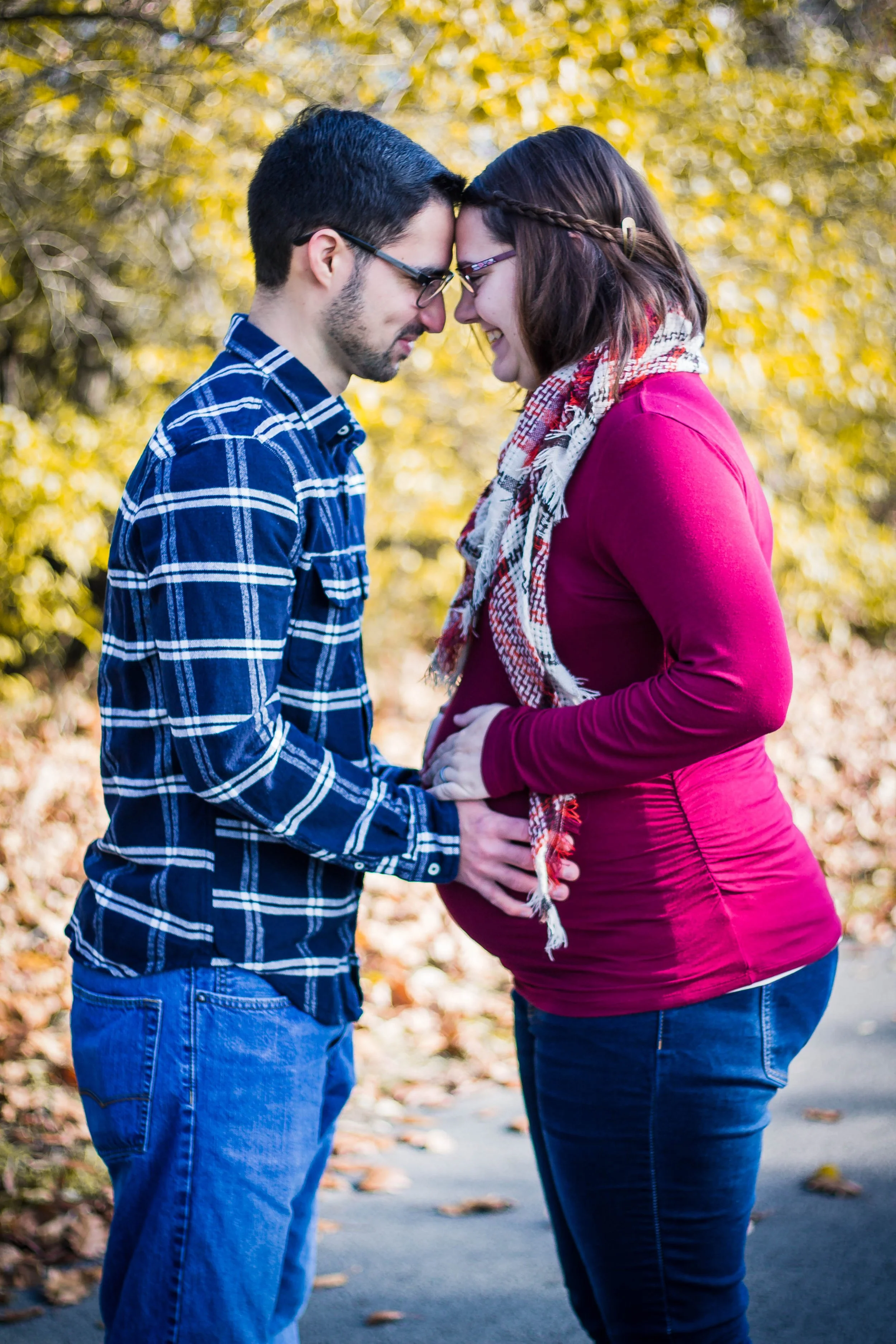 A pregnant woman and a man standing outdoors with their foreheads touching and hands on her belly, surrounded by autumn leaves.