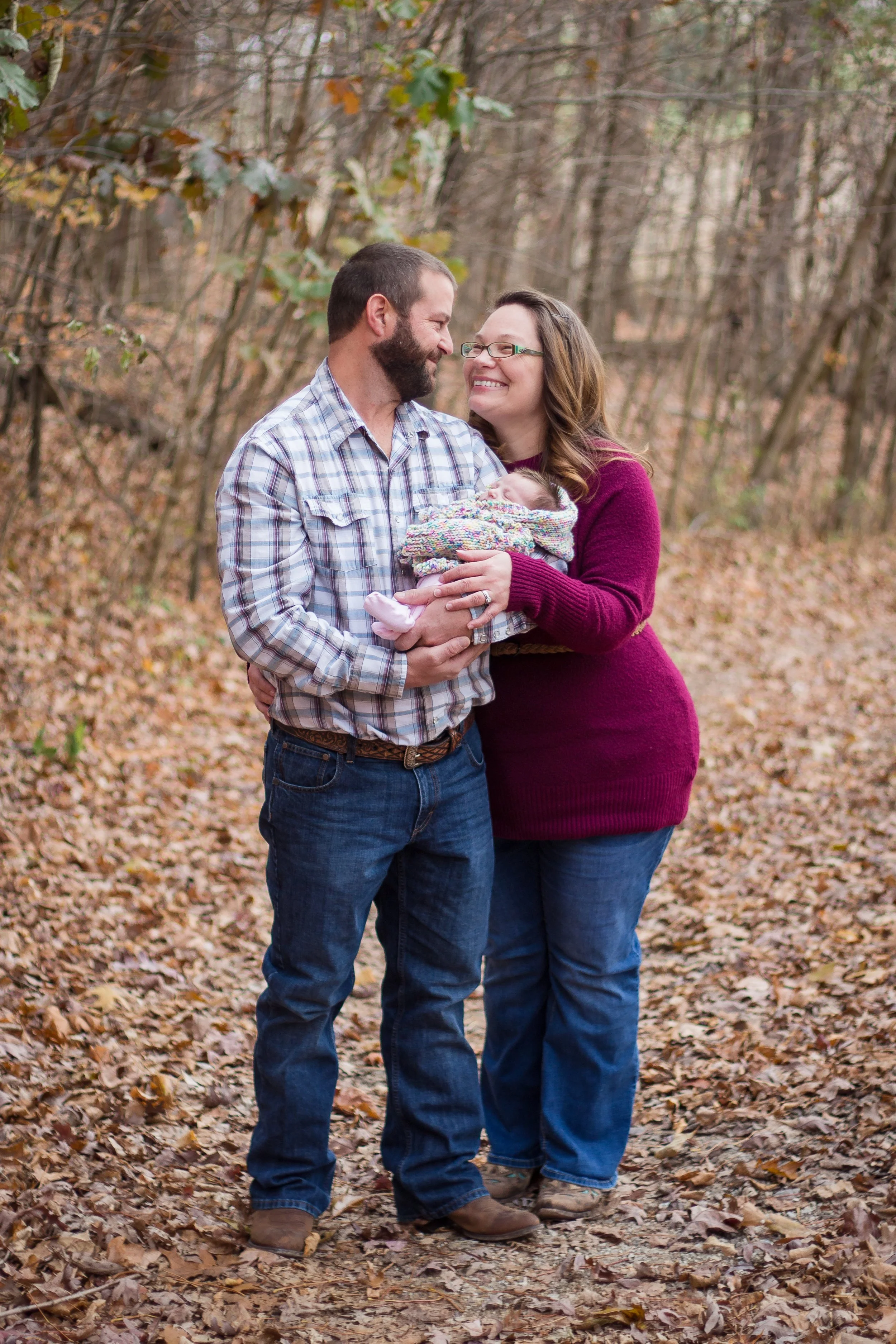 A happy couple standing in a wooded area with fallen autumn leaves, holding their newborn baby wrapped in a colorful blanket and smiling at each other.