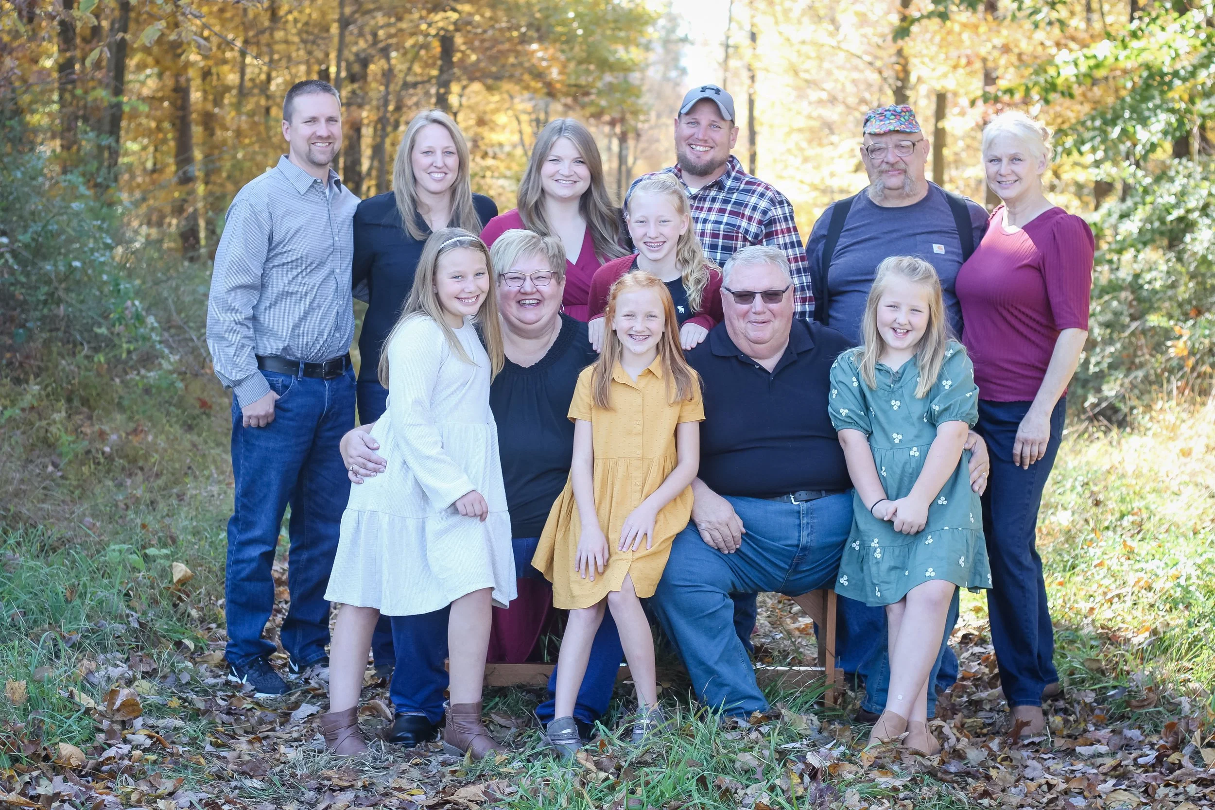 Family group of diverse adults and children outdoors in fall, smiling at camera, surrounded by colorful autumn trees and fallen leaves.