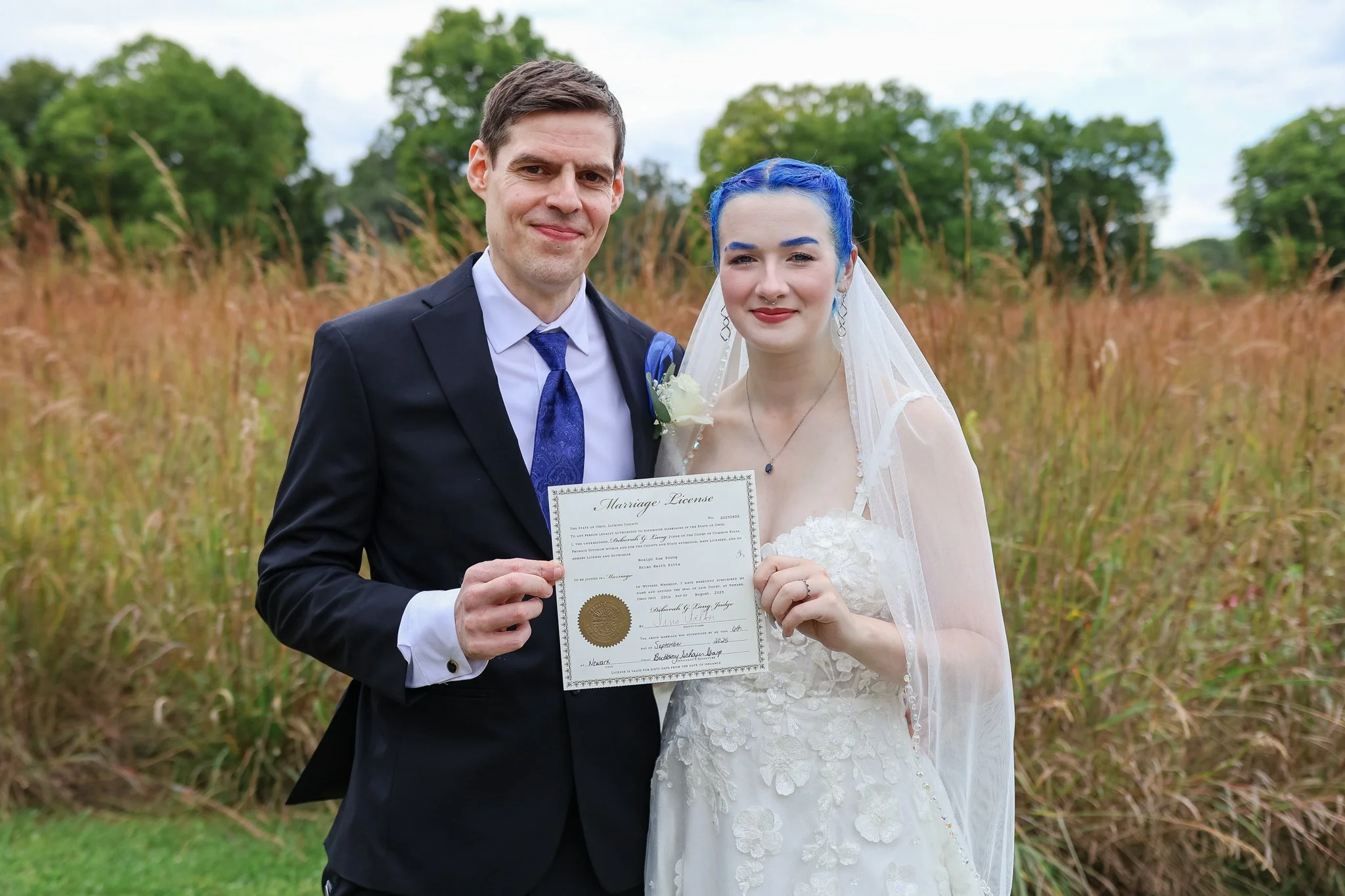 A couple at their wedding, the groom in a black suit and the bride with blue hair and white wedding dress, holding a marriage license certificate outdoors in a grassy field with trees in the background.