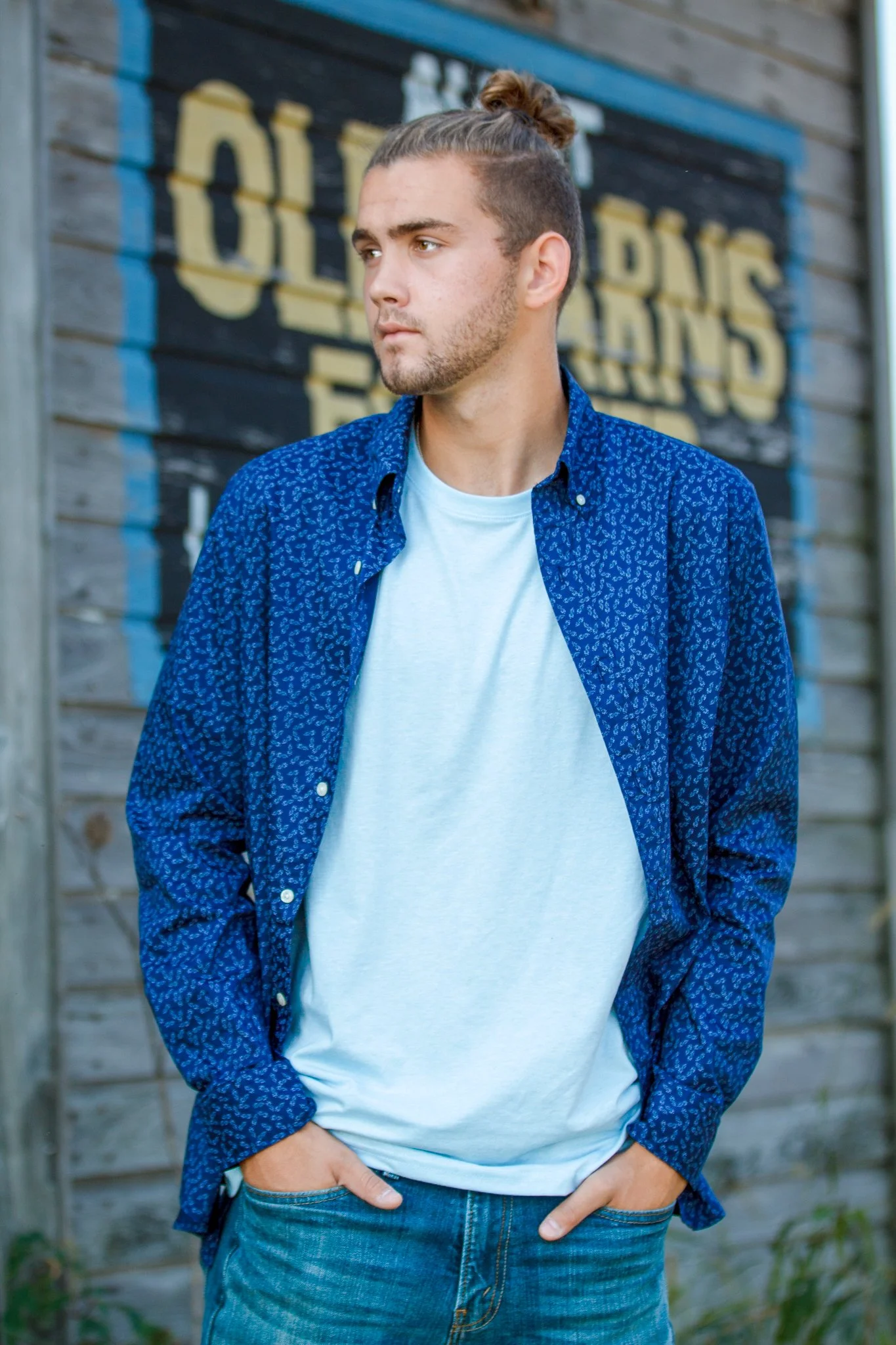 A young man with tied-back hair and a beard, wearing a blue patterned shirt over a white t-shirt, standing outdoors in front of a wooden background with partially visible text.