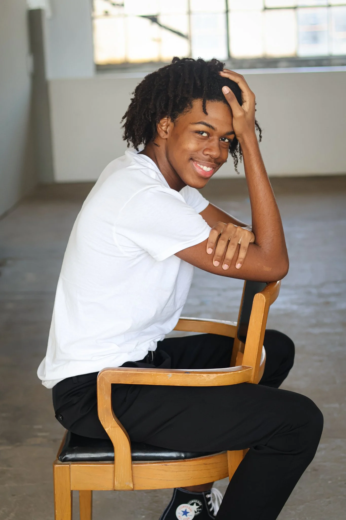 A young man sitting on a wooden chair indoors, smiling, with one hand on his forehead and the other arm resting on the chair's back. He is wearing a white t-shirt, black pants, and black Converse sneakers. The background includes a large window with 