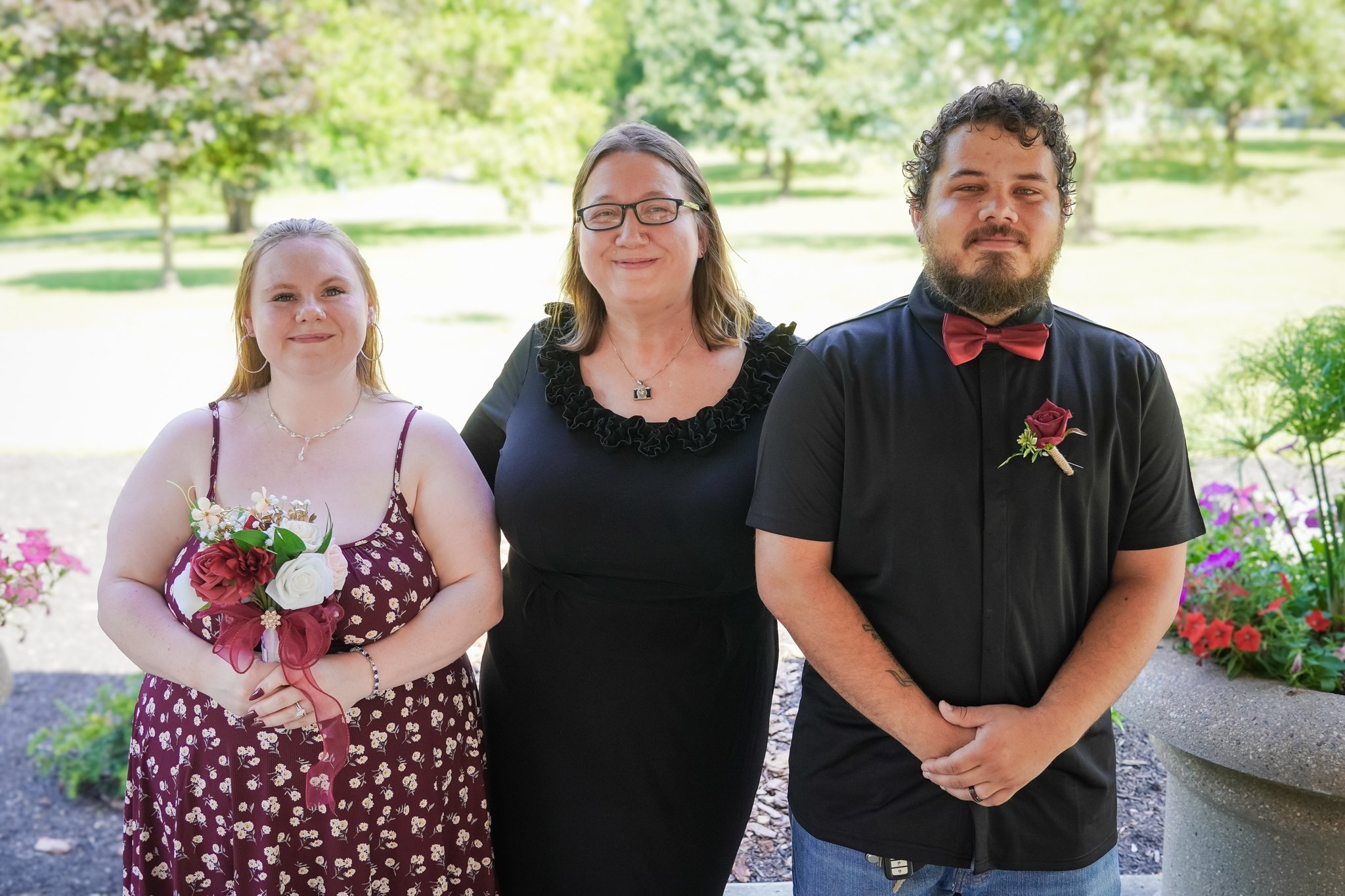 Three people celebrating outdoors, with a woman holding a bouquet, a woman in a black dress with glasses, and a man wearing a tuxedo with a red bow tie and boutonniere, in a park with greenery and flowers.
