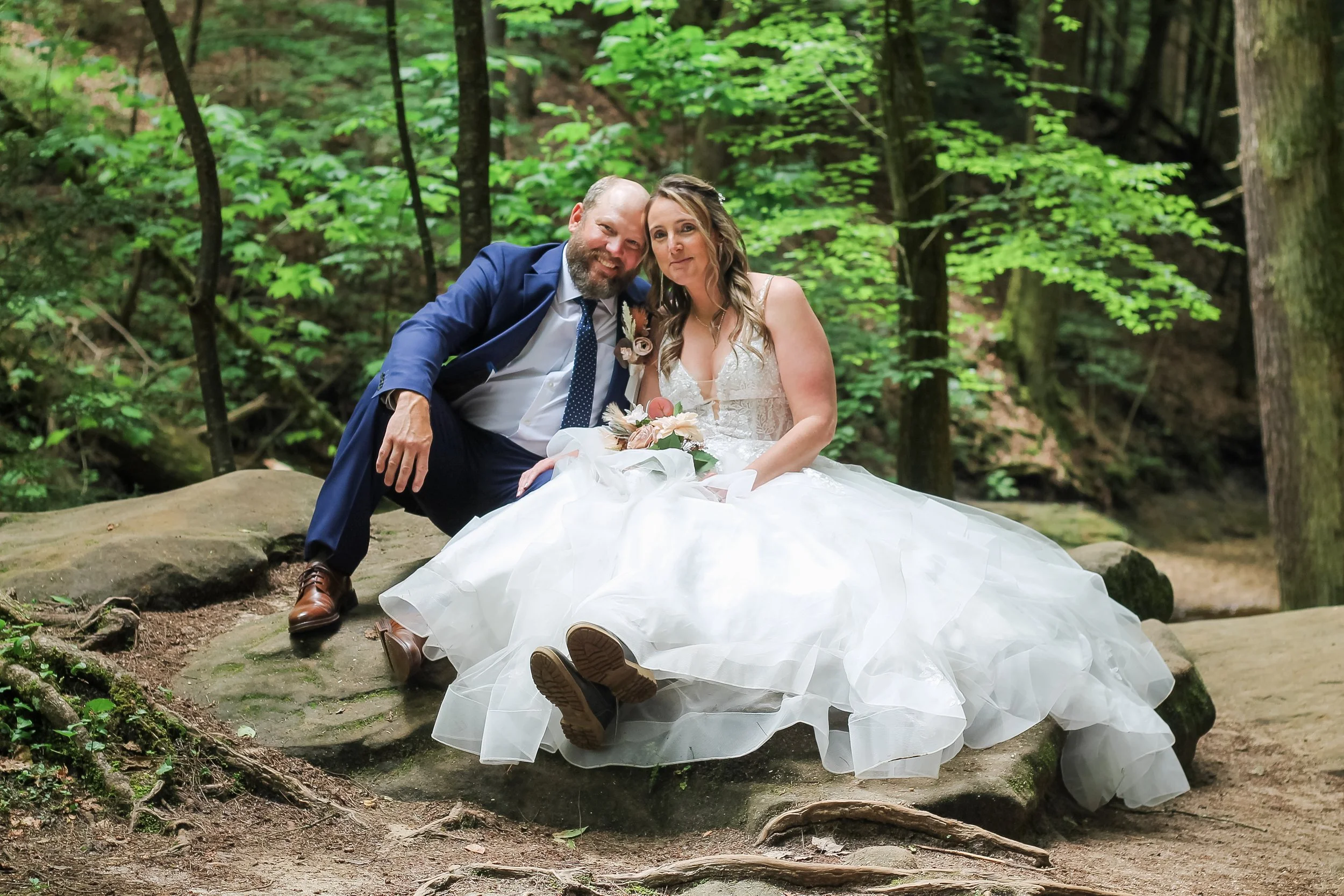 A bride and groom sitting on a rock in a lush green forest. The groom is wearing a blue suit and tie, and the bride is wearing a white wedding dress, holding a bouquet of flowers, both smiling and leaning close to each other.