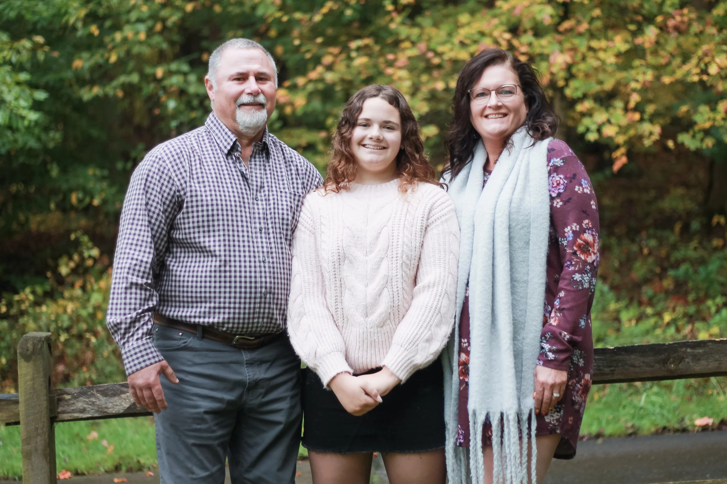 Family of three standing outdoors in a wooded area during fall, smiling at the camera.