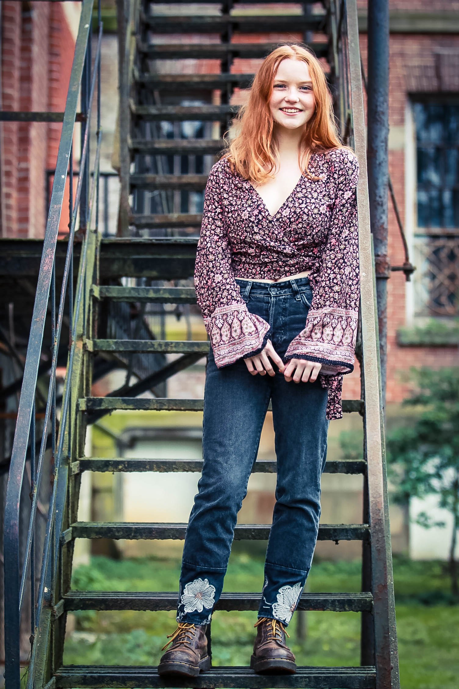 A young woman with long red hair standing on outdoor metal staircase, smiling, wearing a floral blouse, jeans with floral embroidery, and brown lace-up boots, with brick buildings in the background.