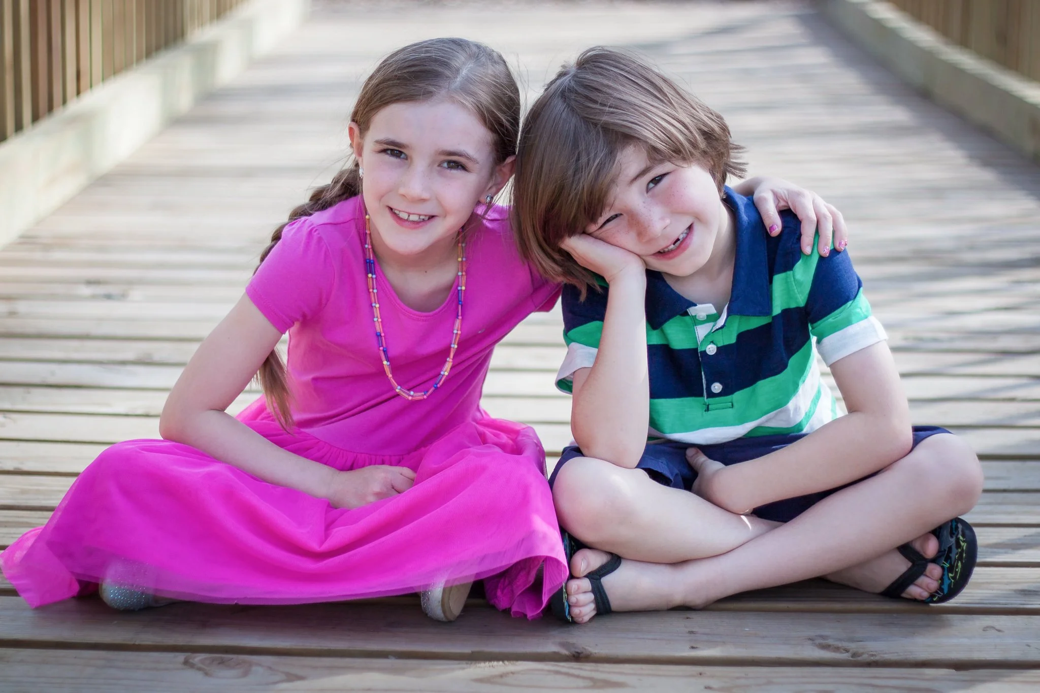 A young girl in a pink dress and a beaded necklace sitting on a wooden dock with a young boy in a striped green and blue shirt. They are smiling and leaning close to each other with the girl resting her arm on the boy's shoulder.