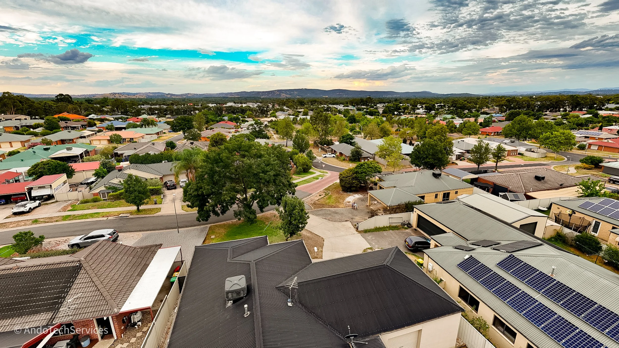 Aerial view of a suburban neighborhood with houses, trees, and streets, under a partly cloudy sky.