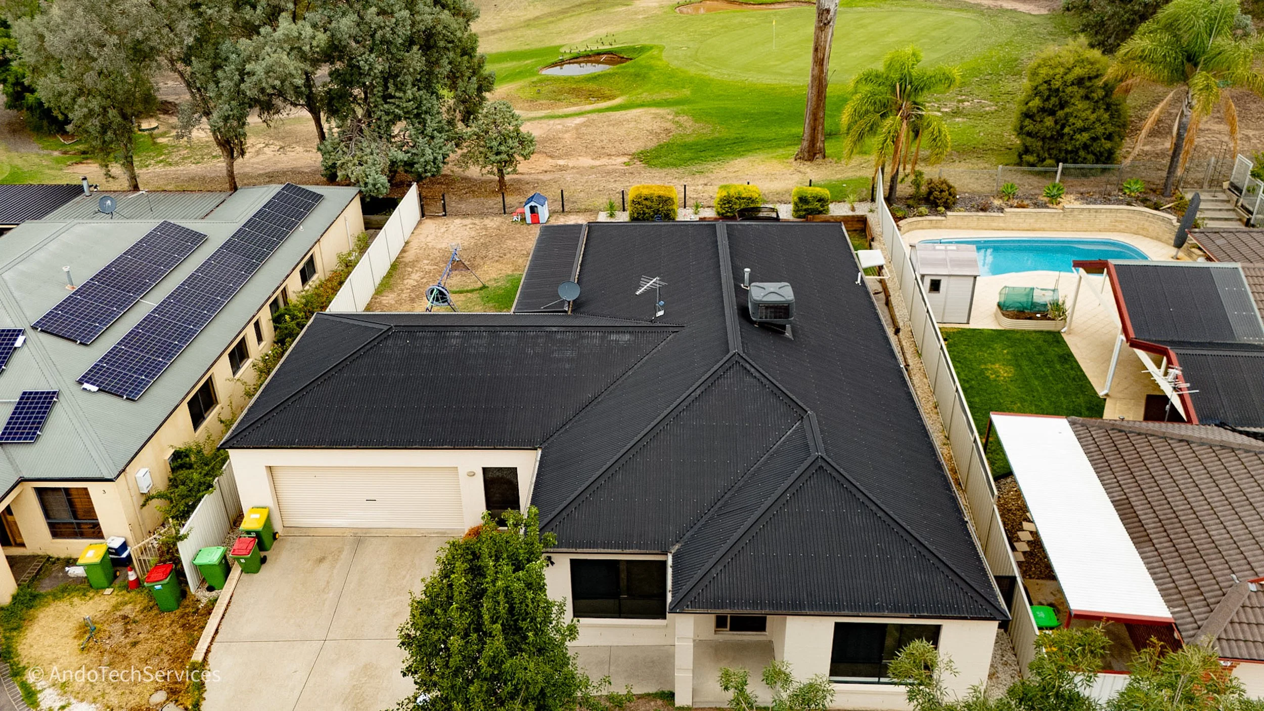 Aerial view of suburban houses, one with solar panels on the roof, next to a green park with trees, a pond, and a golf course in the background, with a backyard pool and outdoor space.
