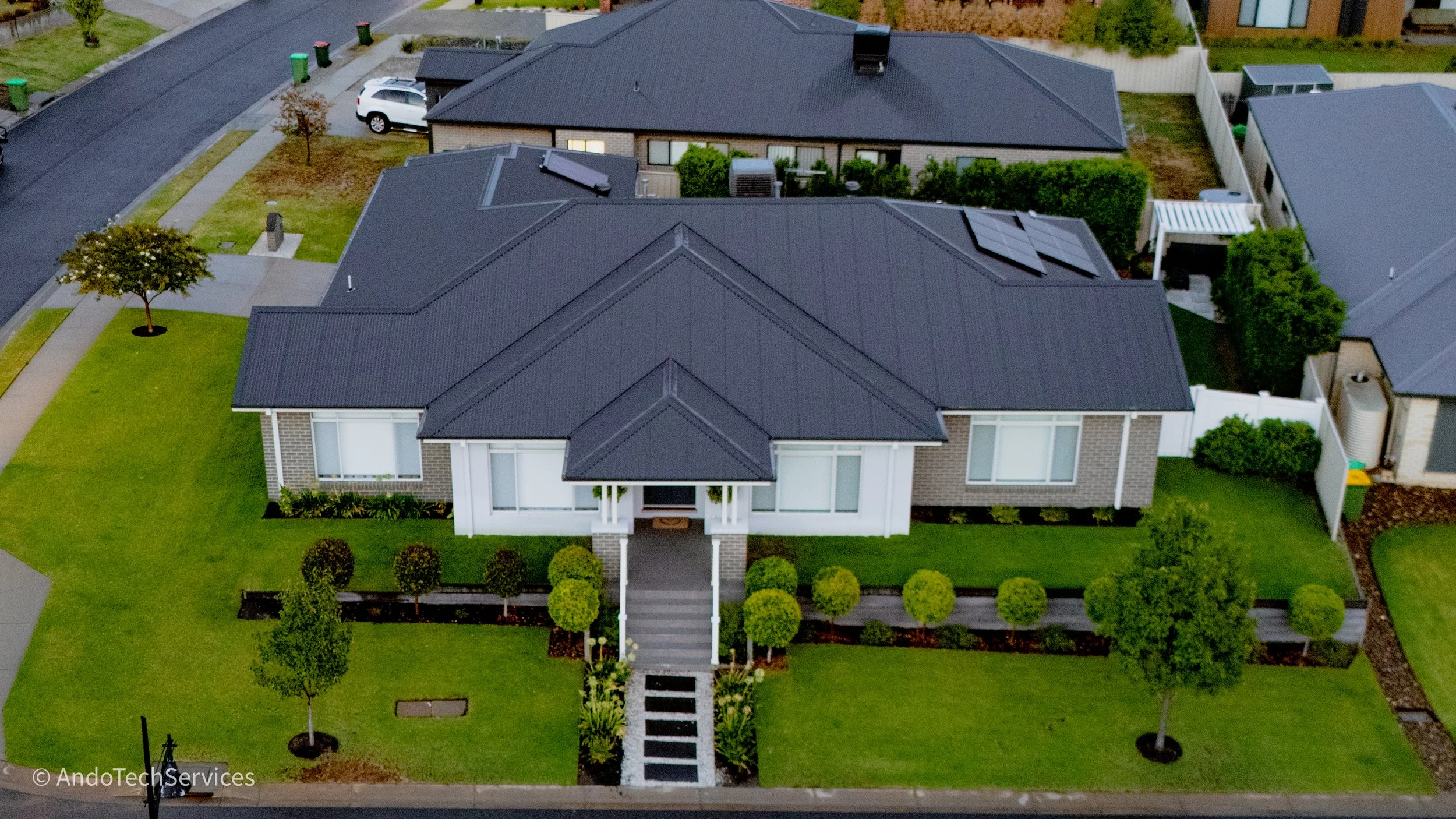 Aerial view of a modern house with a dark roof, white and brick exterior, front steps, evenly spaced bushes, green lawn, and neighboring houses in a suburban neighborhood.