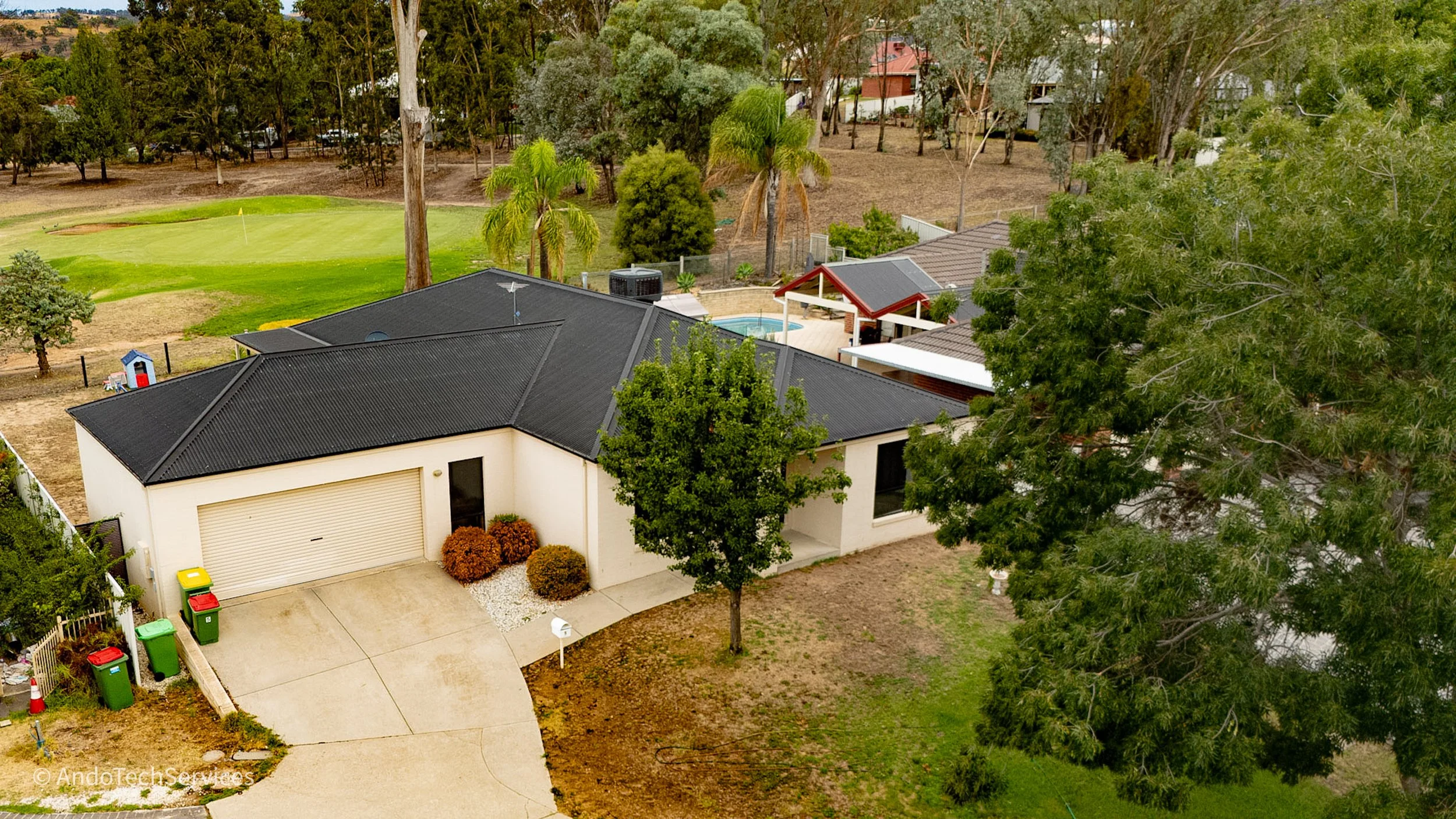 Aerial view of a residential house with a dark roof, surrounded by trees and a yard. There is a small backyard pool, a pool house, and a designated area for waste bins. The house is located next to a green golf course with a putting green and trees.
