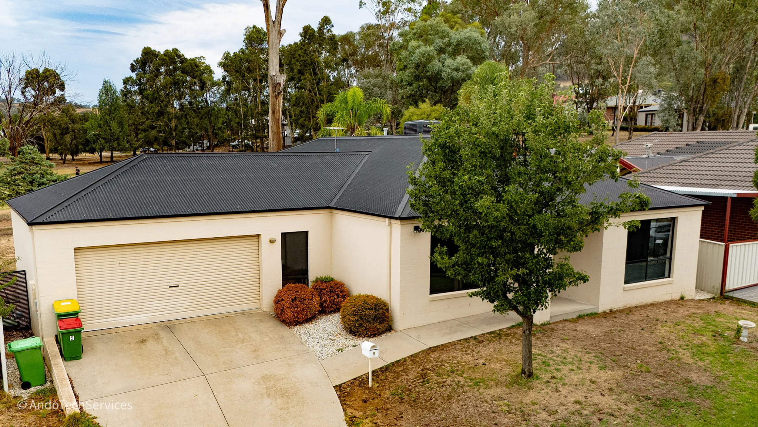 A single-story house with a black metal roof, white exterior walls, a garage door, a large front window, and a small porch. There are three colorful trash bins near the garage, a mailbox, a young tree in the front yard, and some bushes with autumn le