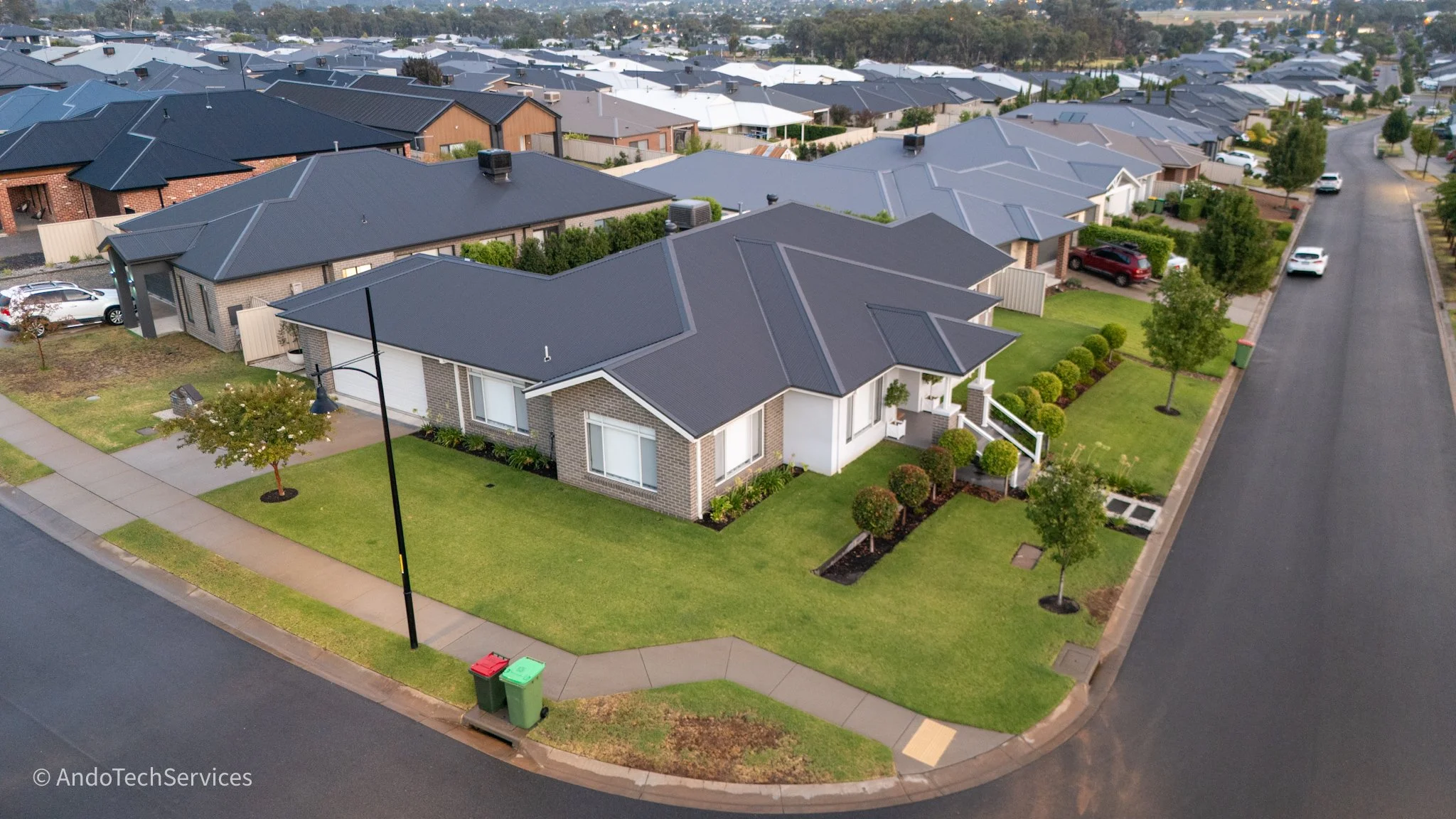 An aerial view of a suburban neighborhood showing single-family homes with driveways, well-maintained lawns, trees, and parked cars along the street.