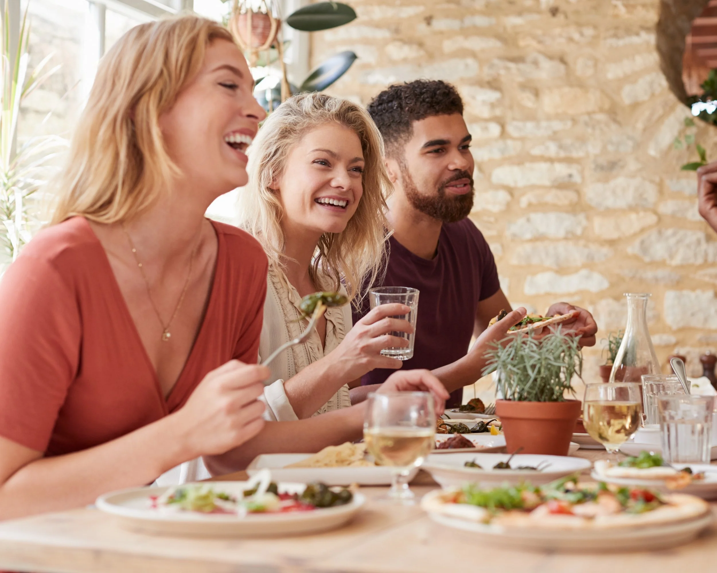 Three friends enjoying a meal together at a restaurant, with food and drinks on the table and a brick wall background.