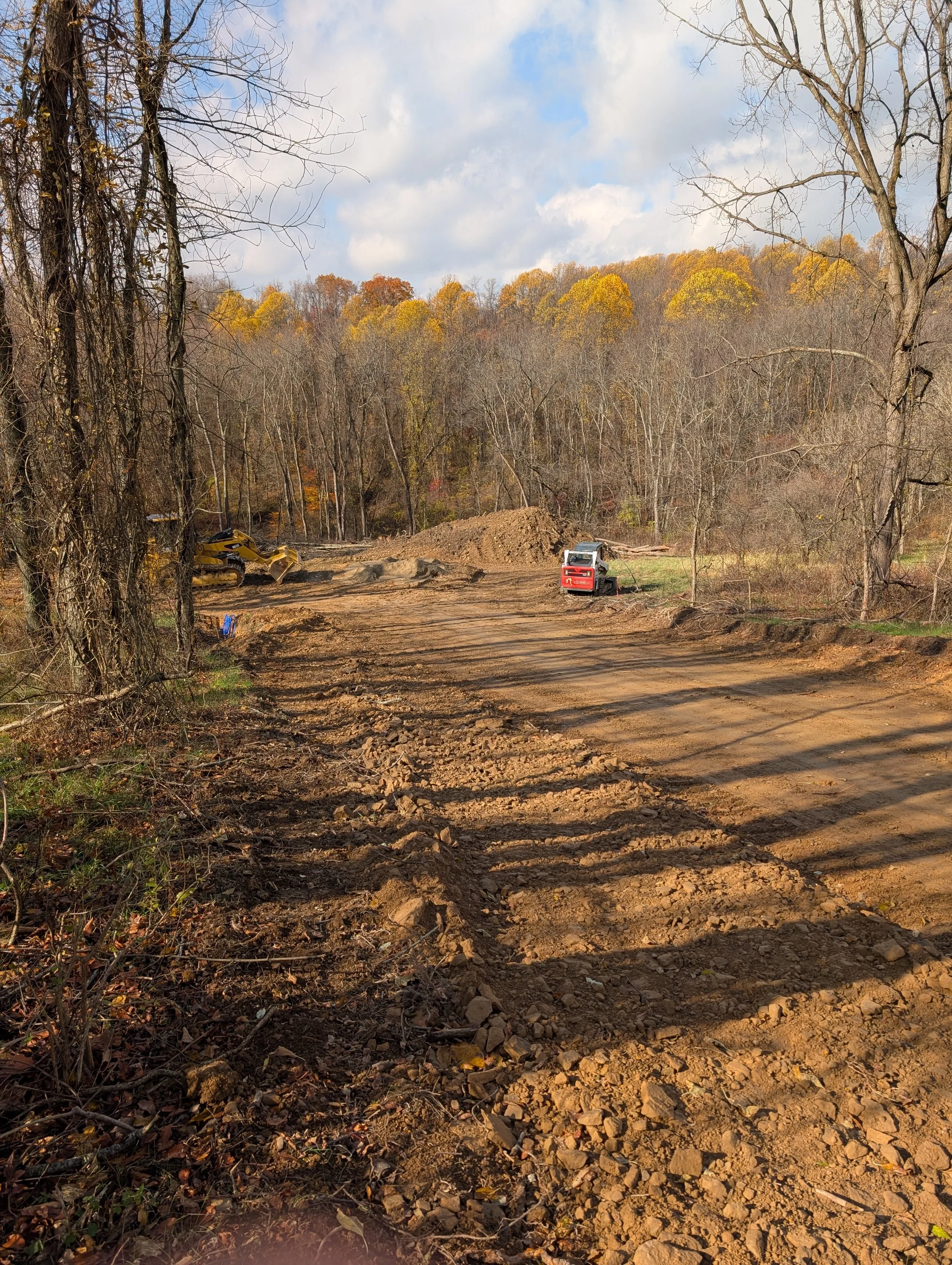 A dirt road in a forested area with some construction equipment and a small bulldozer, autumn-colored trees in the background, and partly cloudy sky.