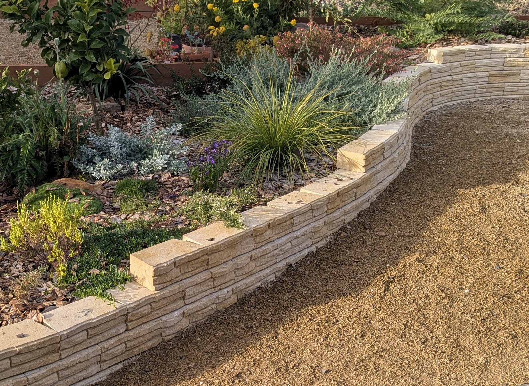 A professionally designed, built, and landscaped garden bed with various green plants and flowers, bordered by a low stacked stone wall, with a gravel area beside it.