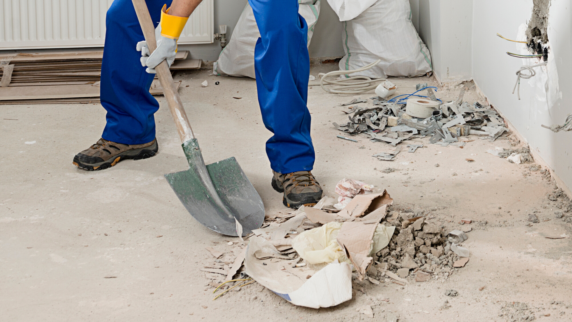 Worker in blue work pants and boots using a shovel to remove debris from a construction site. Dust and scattered materials on the floor with exposed wiring and a wall with holes.