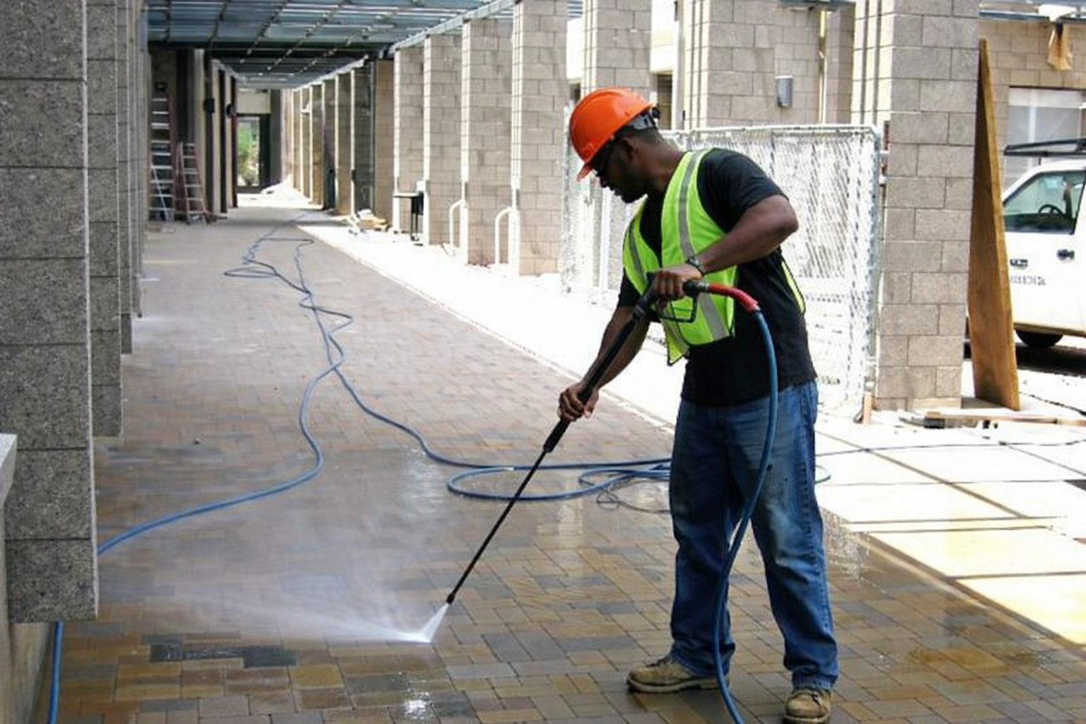 A man wearing a safety vest, helmet, and shoes is pressure washing a brick sidewalk outdoors near a building construction site.