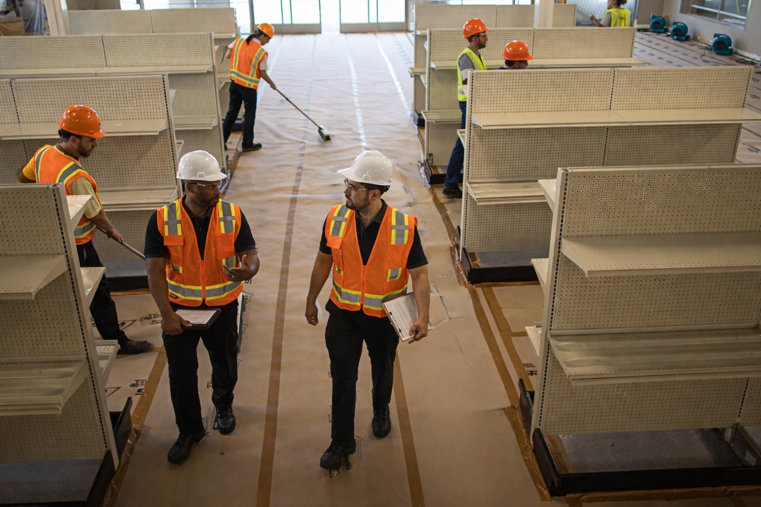 Construction workers wearing safety vests and helmets inspect empty retail shelving units inside a building under construction, with workers cleaning the floor in the background.