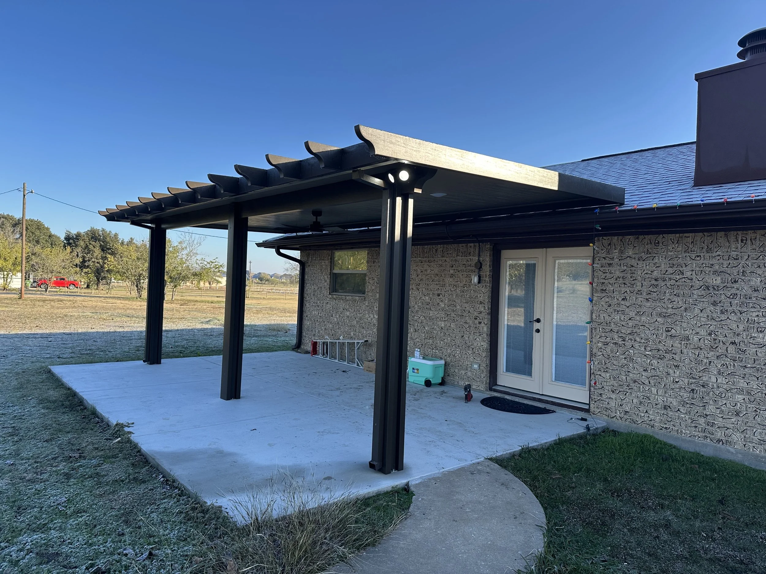Back patio with a concrete floor and black pergola structure. French doors with glass panels lead into the house, which has stone exterior walls. A small cool box, ladder, and some tools are on the patio. The yard is open with trees and a red vehicle in the distance under a clear blue sky.