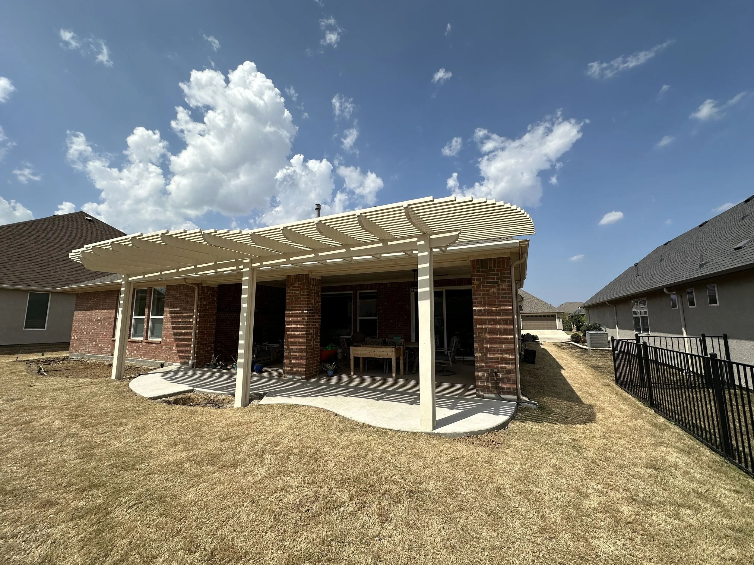 Backyard patio with white pergola, brick house, and neighboring houses under a partly cloudy sky.
