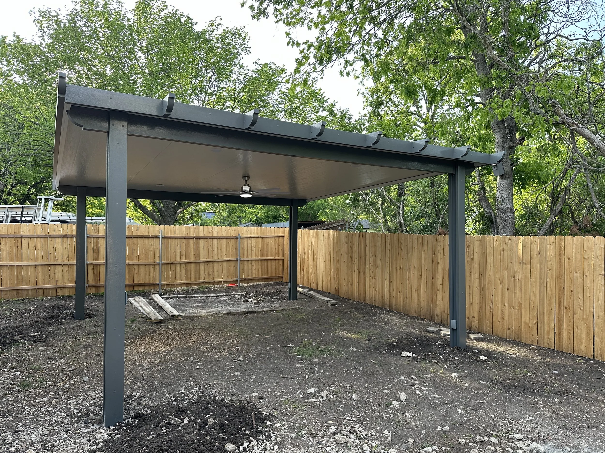 Empty backyard with newly installed metal carport, surrounded by a wooden privacy fence, and trees in the background.