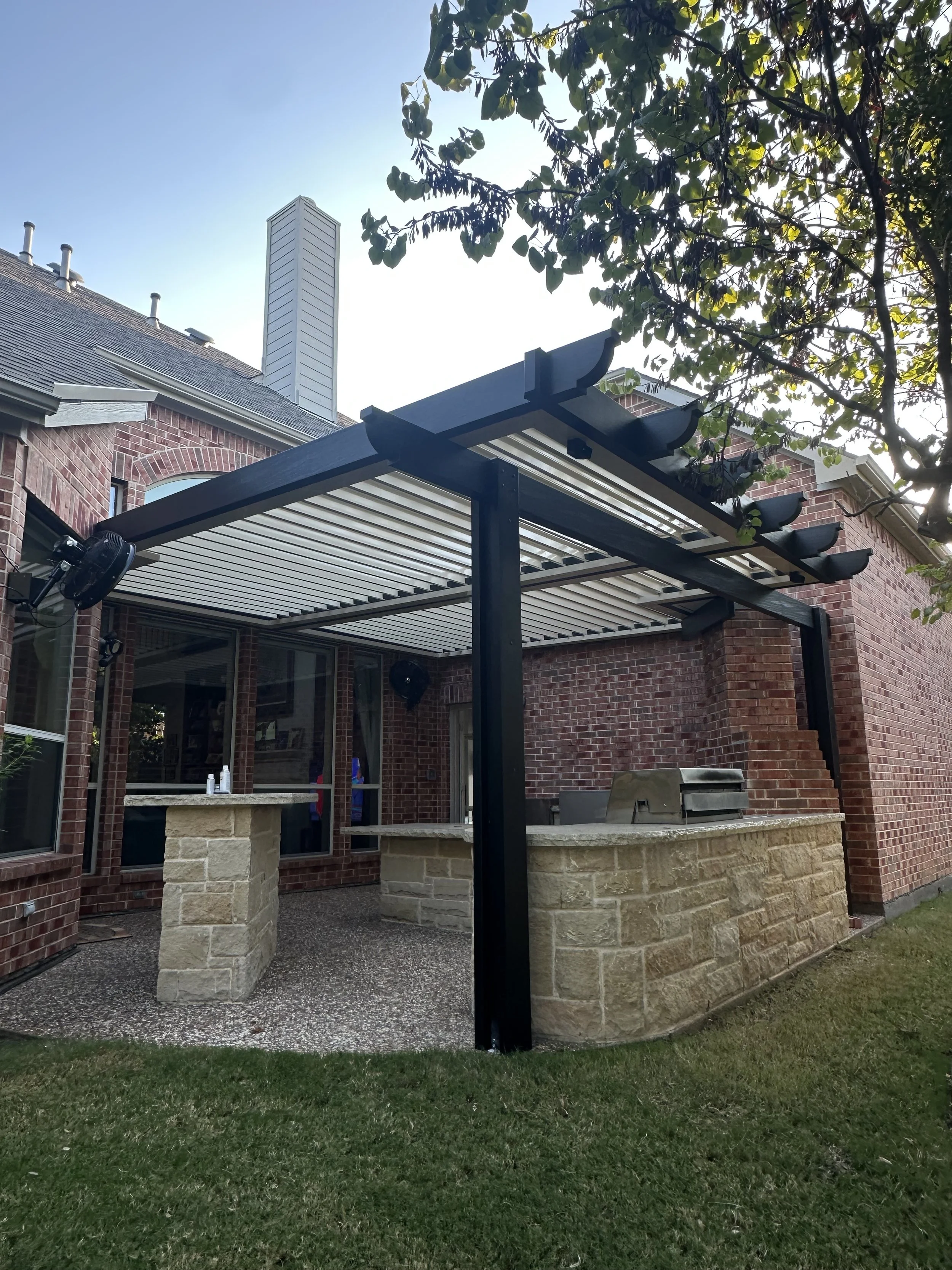 A backyard patio with a black metal pergola, brick and stone outdoor kitchen, and a barbecue grill.