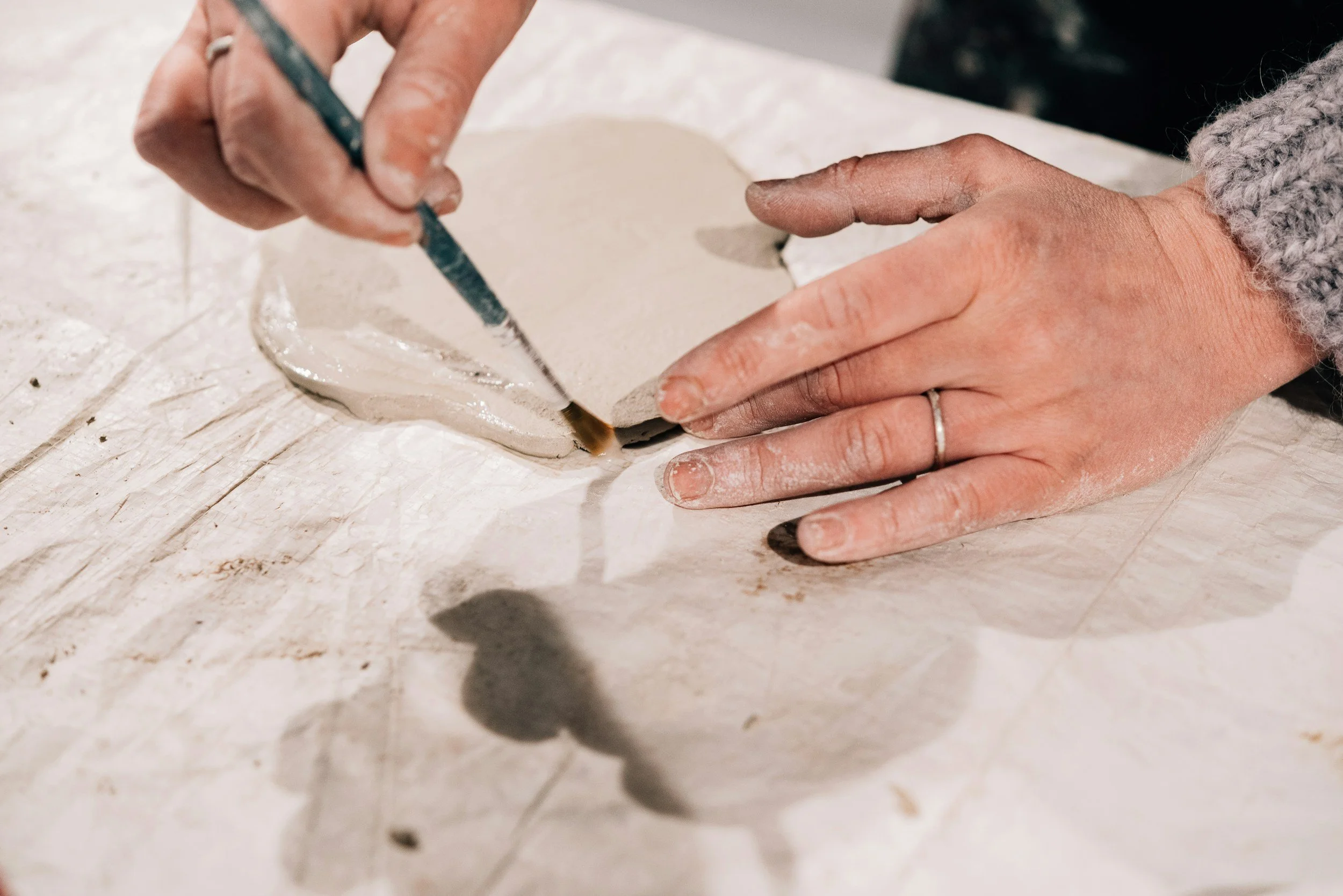 Someone working on a ceramic piece using a small brush, with their hands covered in dust or clay, on a work surface.