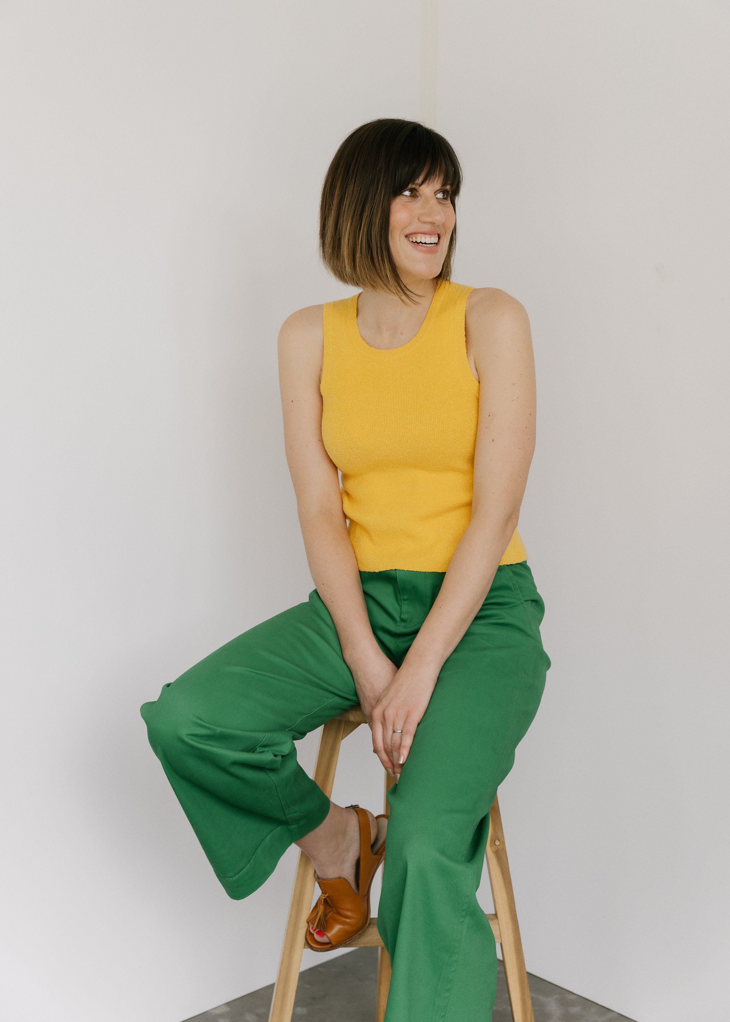 A woman sitting on a wooden stool, looking to her left, smiling, wearing a yellow sleeveless top, green pants, and brown high-heeled shoes, against a plain white background.