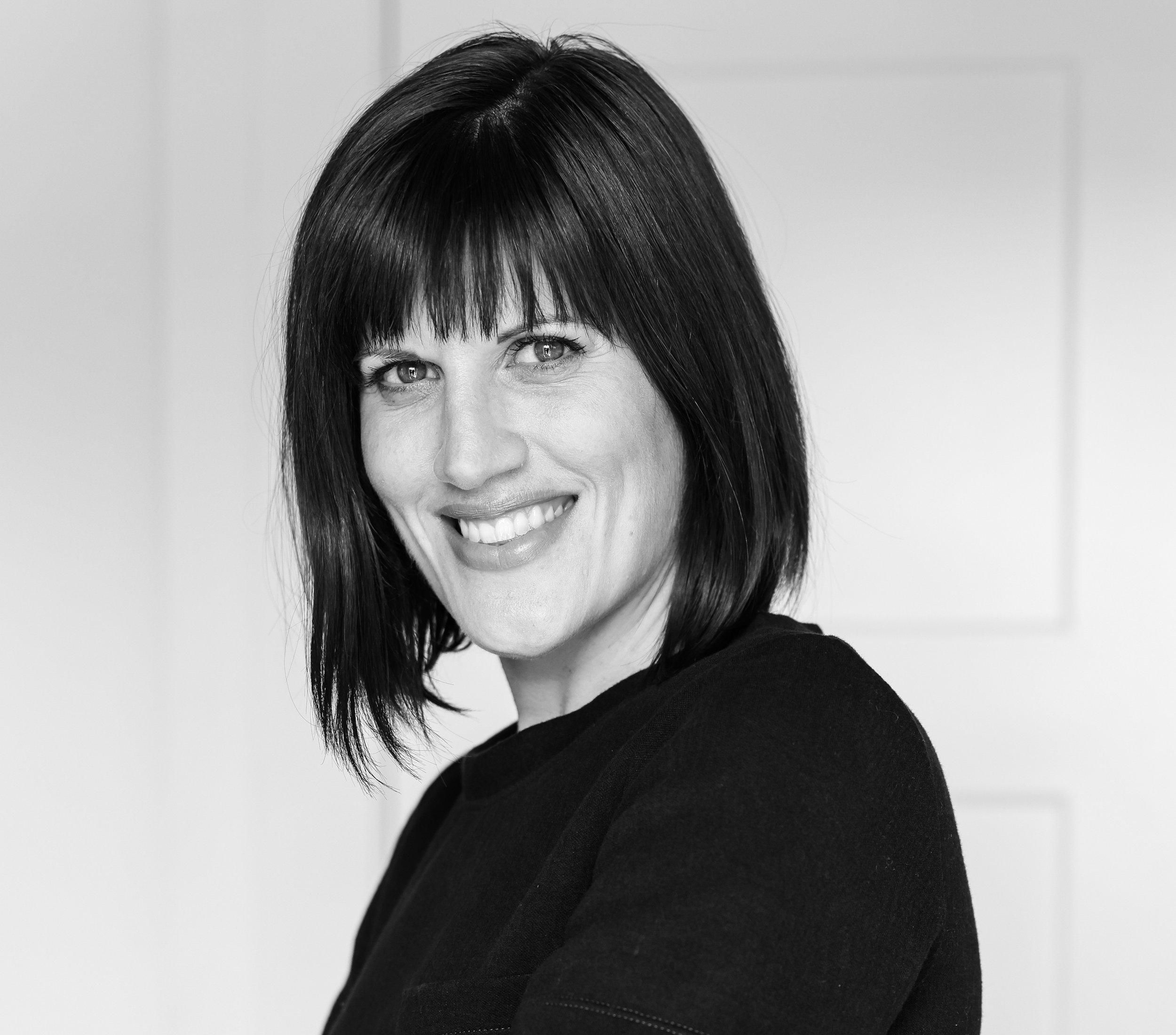 Black and white portrait of a woman with shoulder-length dark hair, smiling and wearing a black top, standing in front of a plain light-colored background.