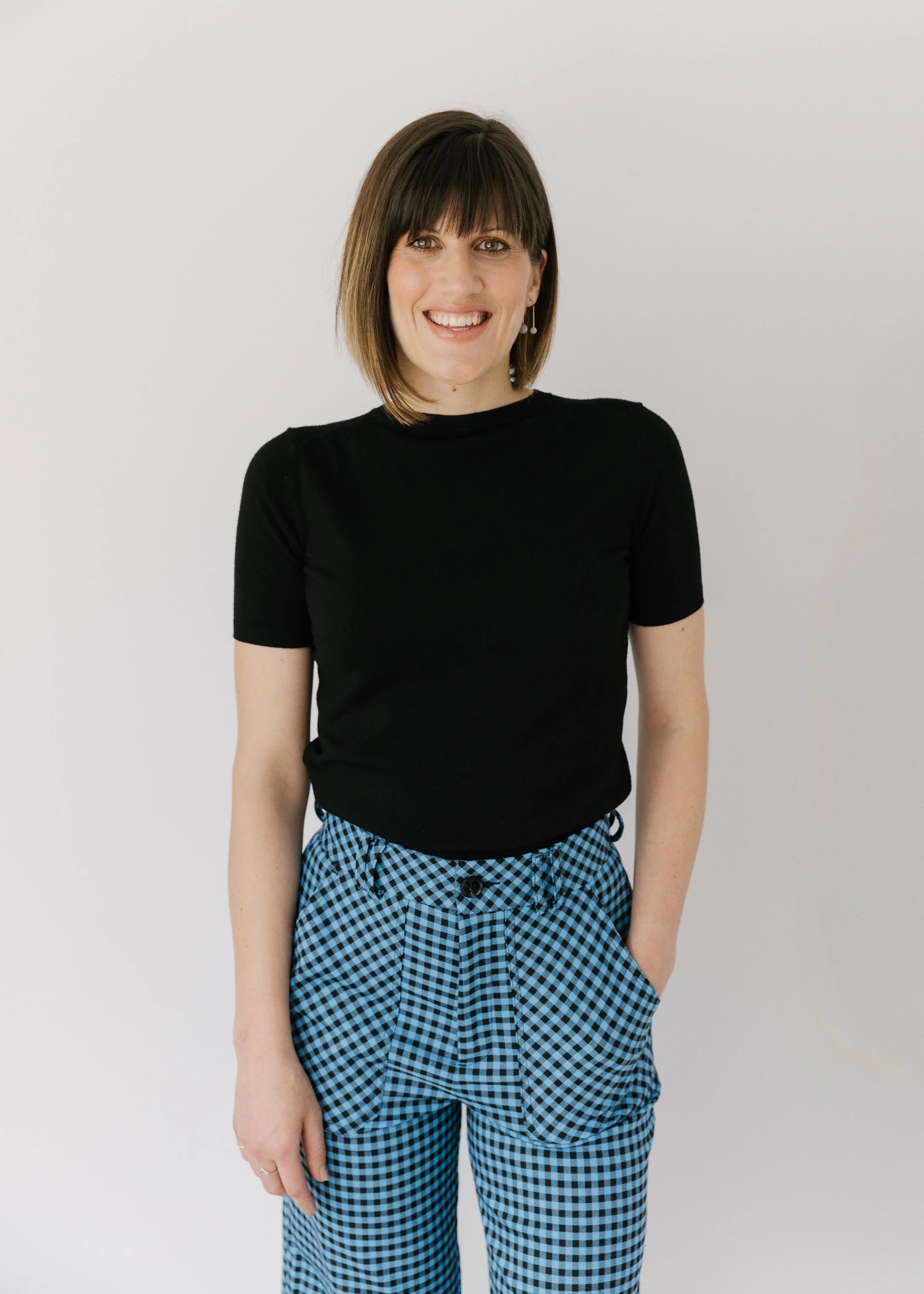 A woman with shoulder-length brown hair, wearing a black short-sleeve top and blue and black gingham pants, smiling and standing against a plain white background.