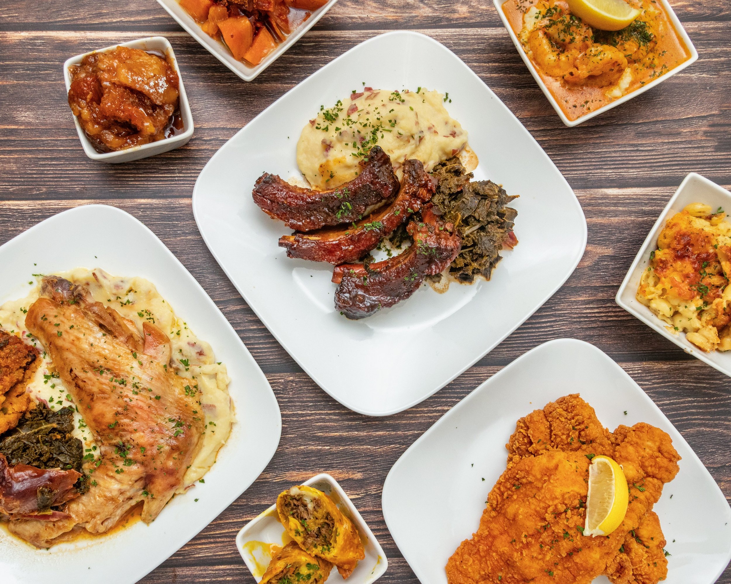 Various Indian dishes on white plates and bowls on a wooden table, including chicken curry, lamb ribs, fried chicken with lemon, mashed potatoes with gravy, vegetable curry, and small bowls of chutney and vegetable preparations.