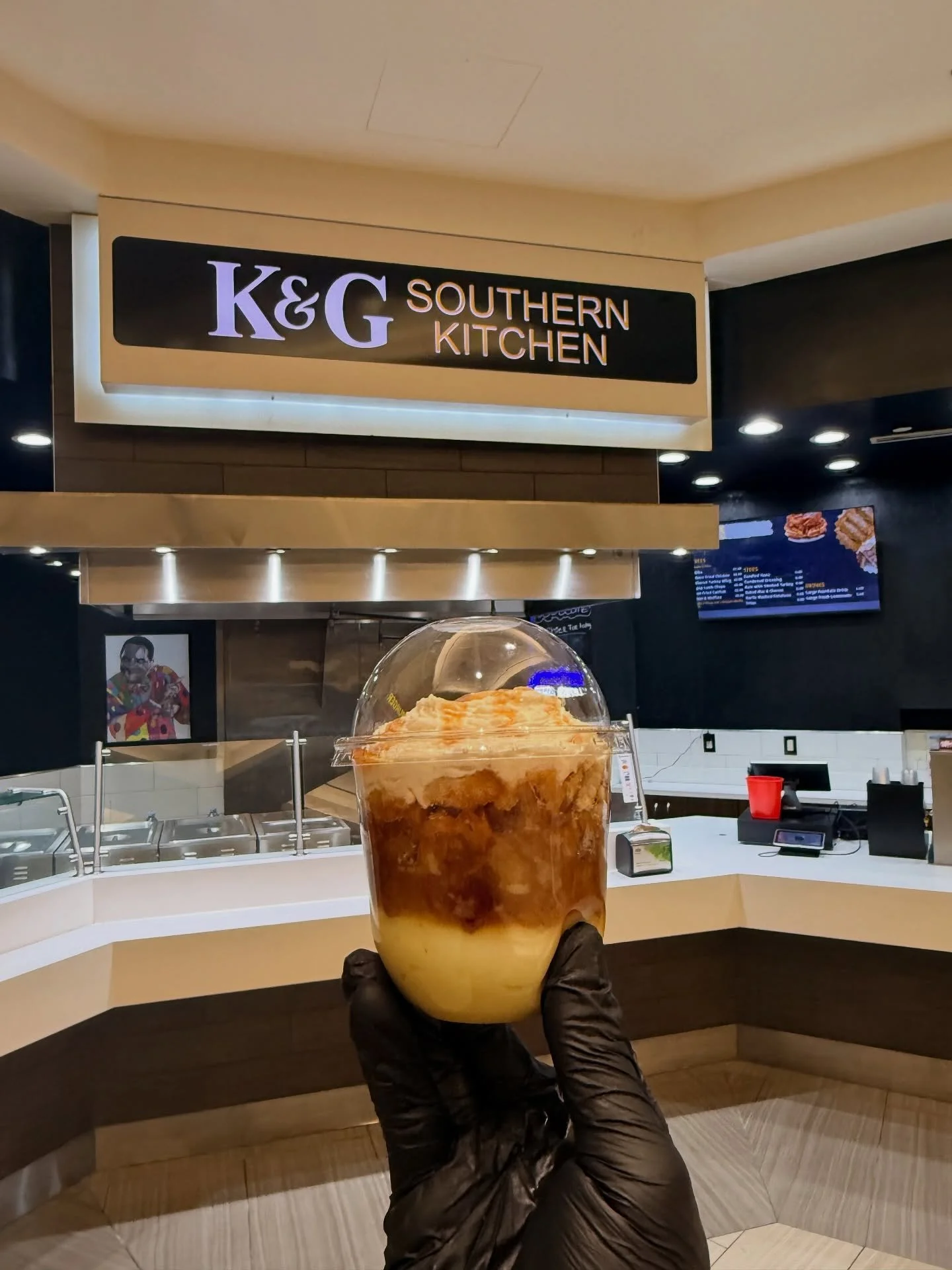 A hand in a black glove holding a cup of coffee with milk and caramel syrup at a K&G Southern Kitchen restaurant counter.