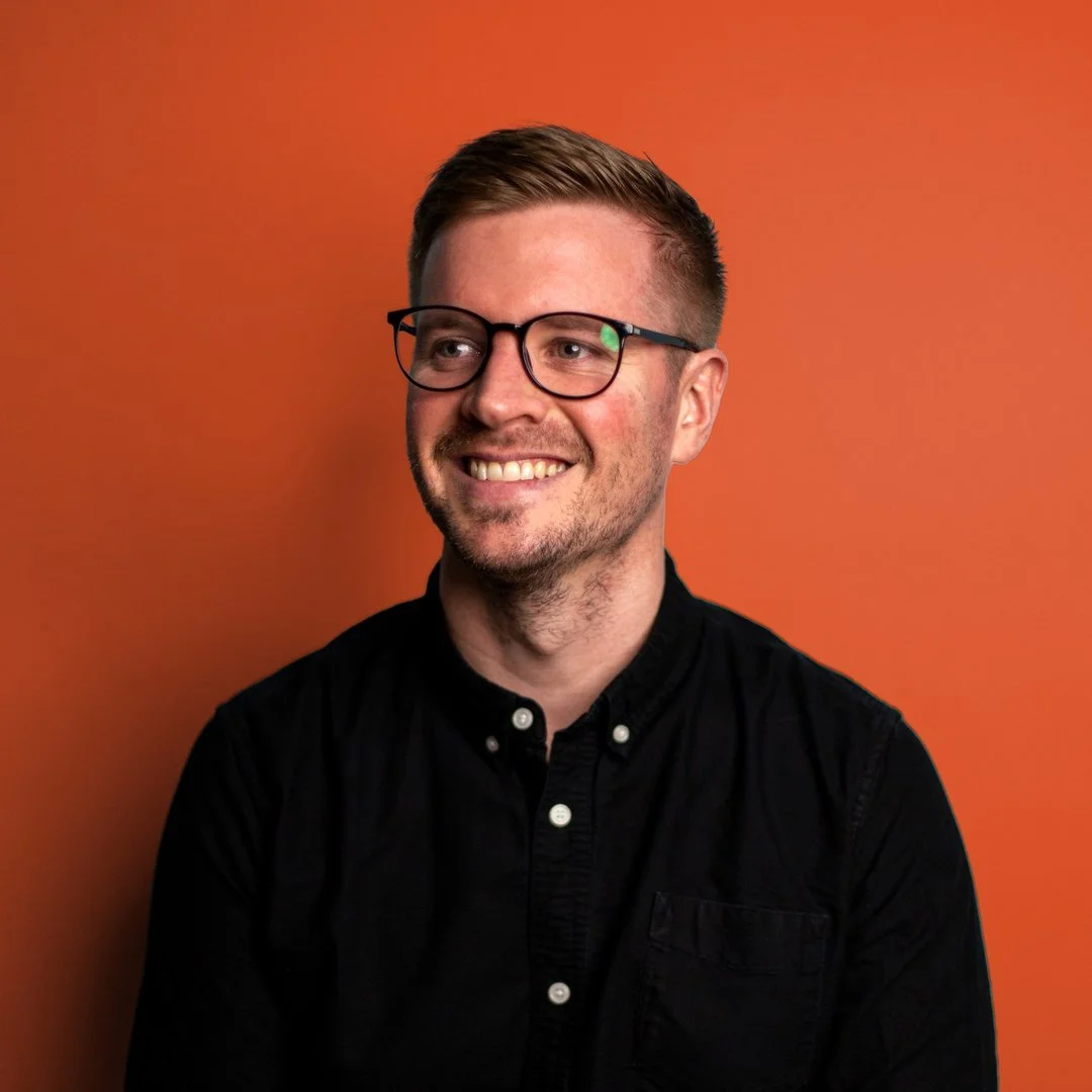 Photo of Shane smiling, looking away from the camera. He has short light-brown hear, wears dark-rimmed glasses, and a black button-up shirt.