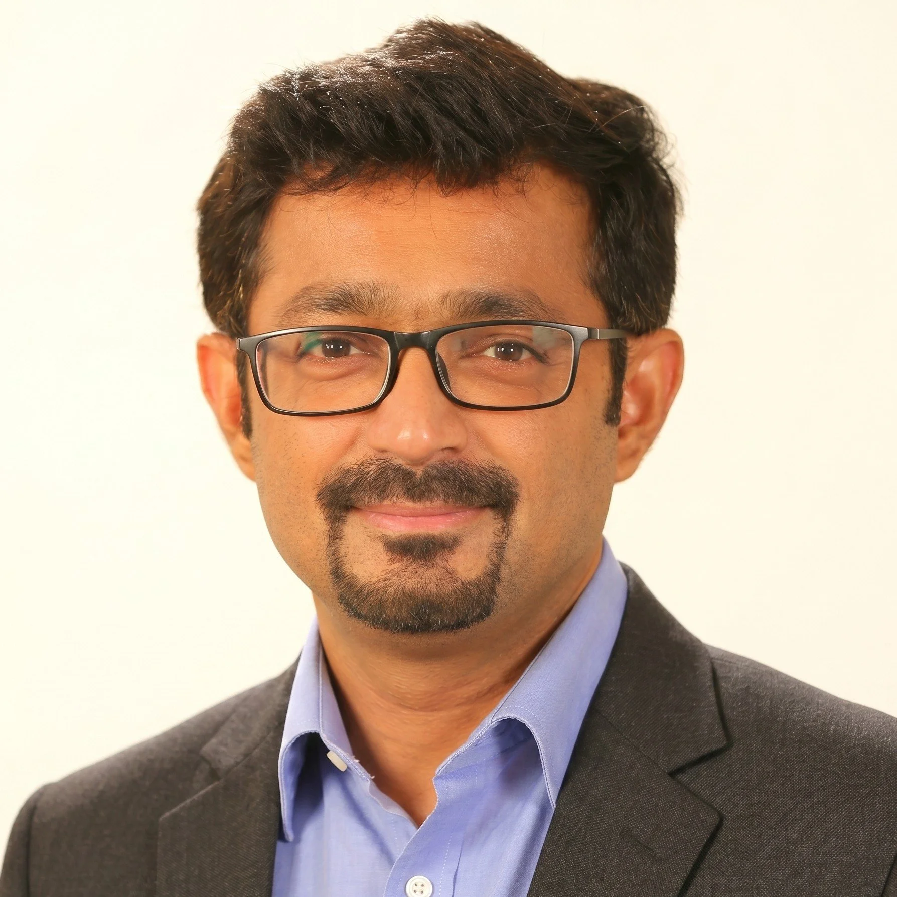 Photo of Subho, smiling at the camera. He has short dark hair and goatee. He wears dark rimmed glasses, and a grey blazer over a blue collared shirt.