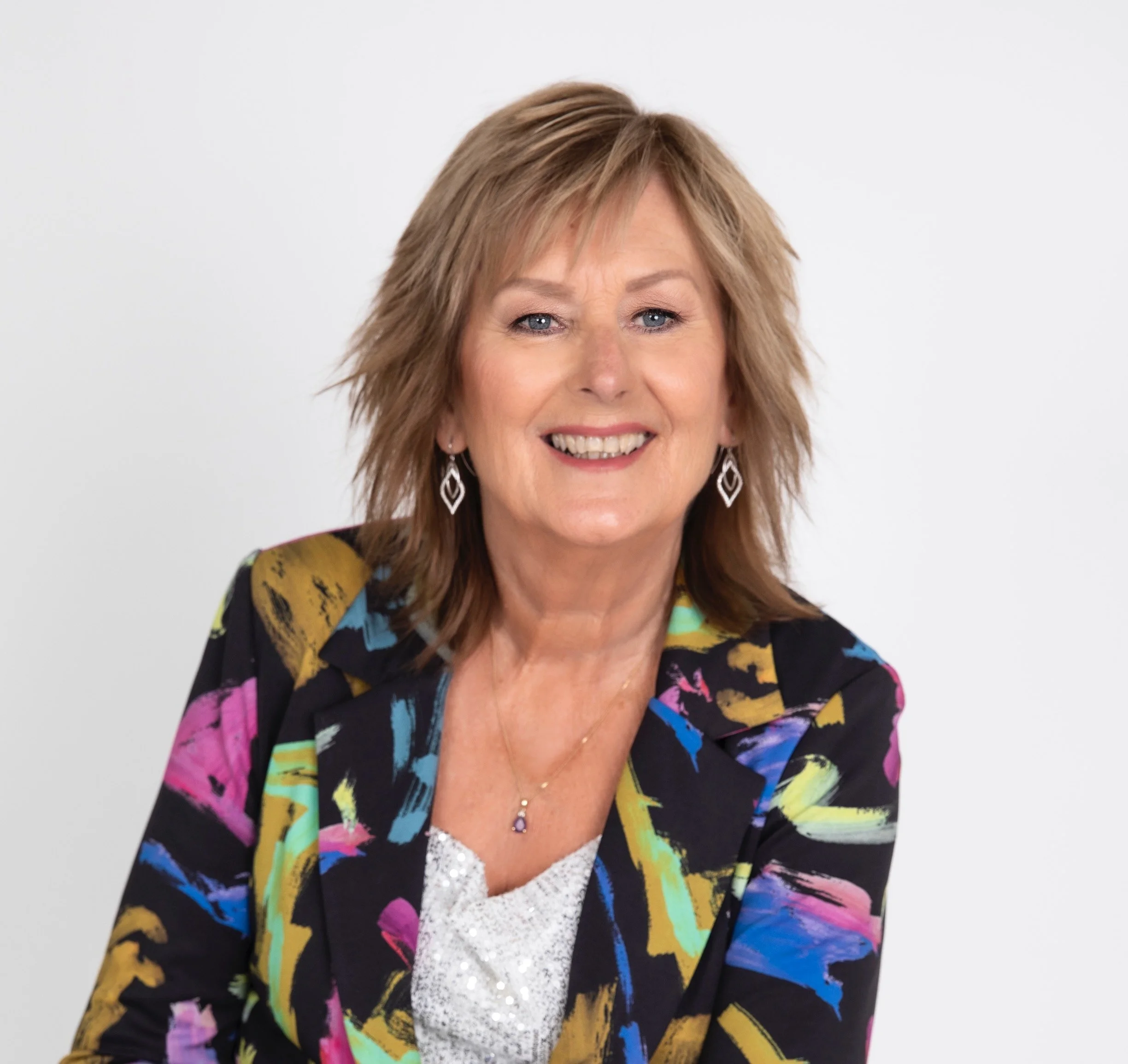 Photo of Neryl smiling at the camera. She has dark blonde shoulder-length hair, and wears a bright-coloured blazer over a white shirt.