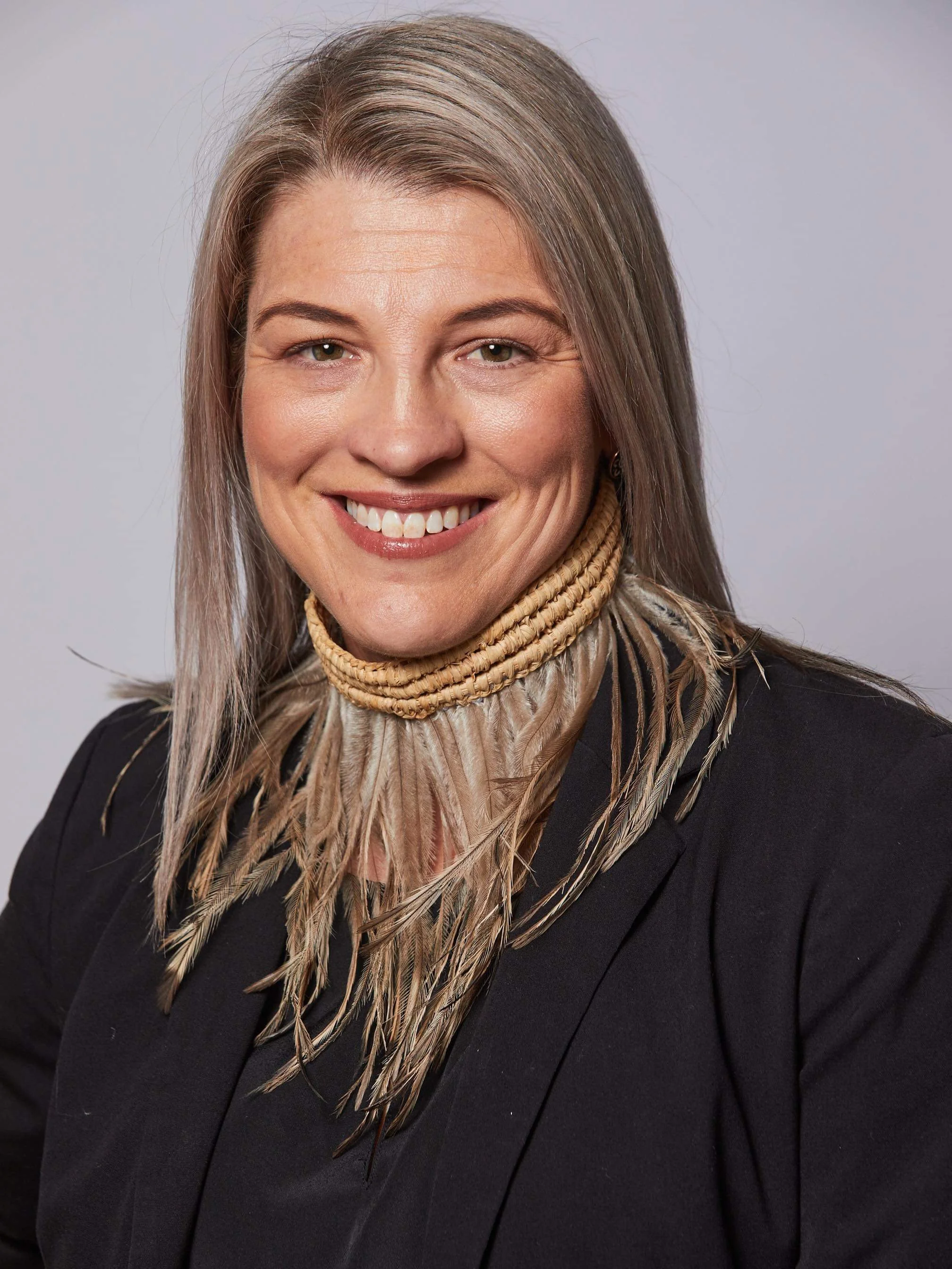 Photo of Tammy smiling at the camera. She has long straight grey hair, and wears a traditional feathered necklace over a dark blazer.