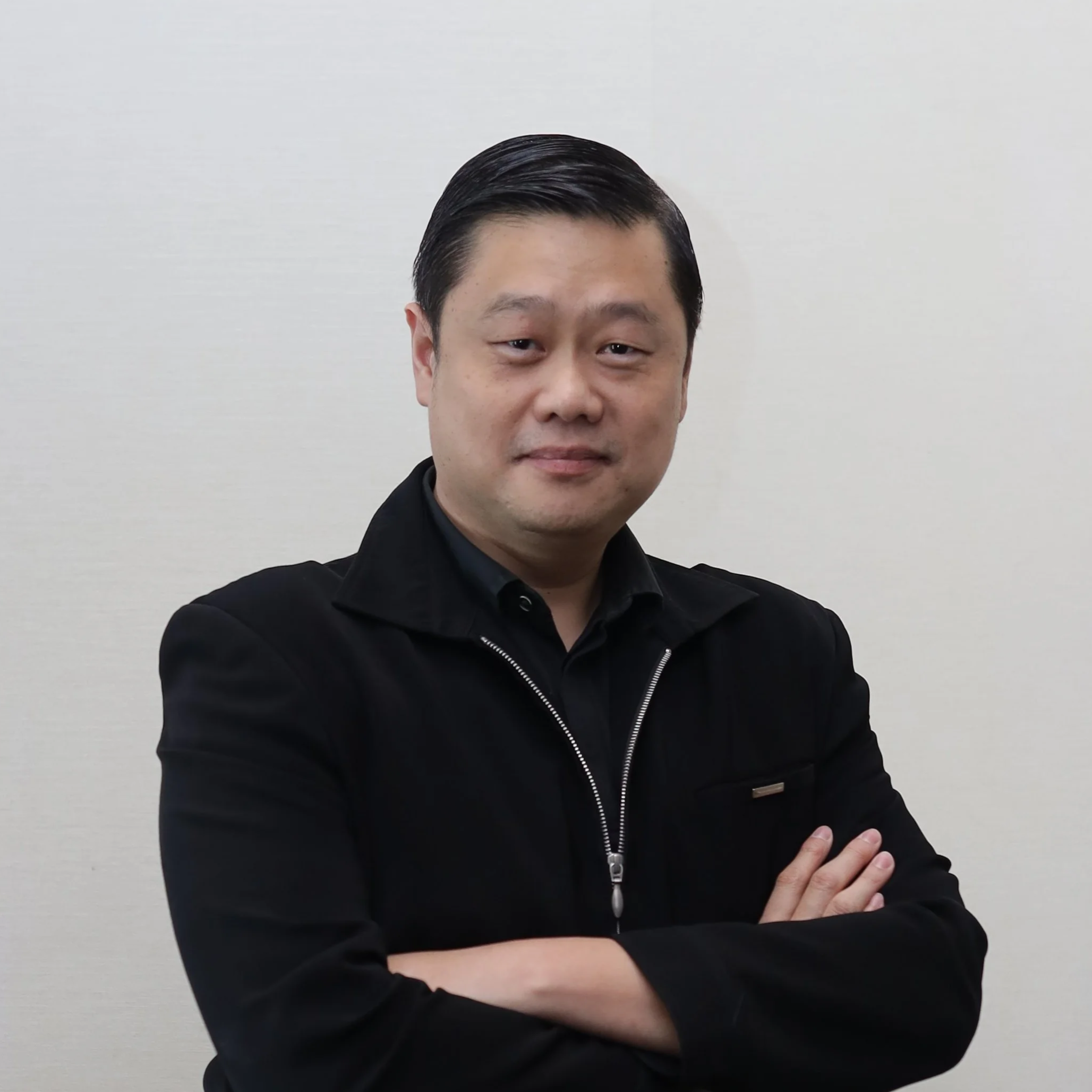 Photo of Dr Lim smiling at the camera. He has short dark hair and stands with his arms folded. He wears a black zip-up collared jacket over a black collared shirt.