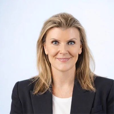 Photo of Kath smiling at the camera. She has shoulder-length dark blonde hair, and wears a dark blazer over a white shirt.