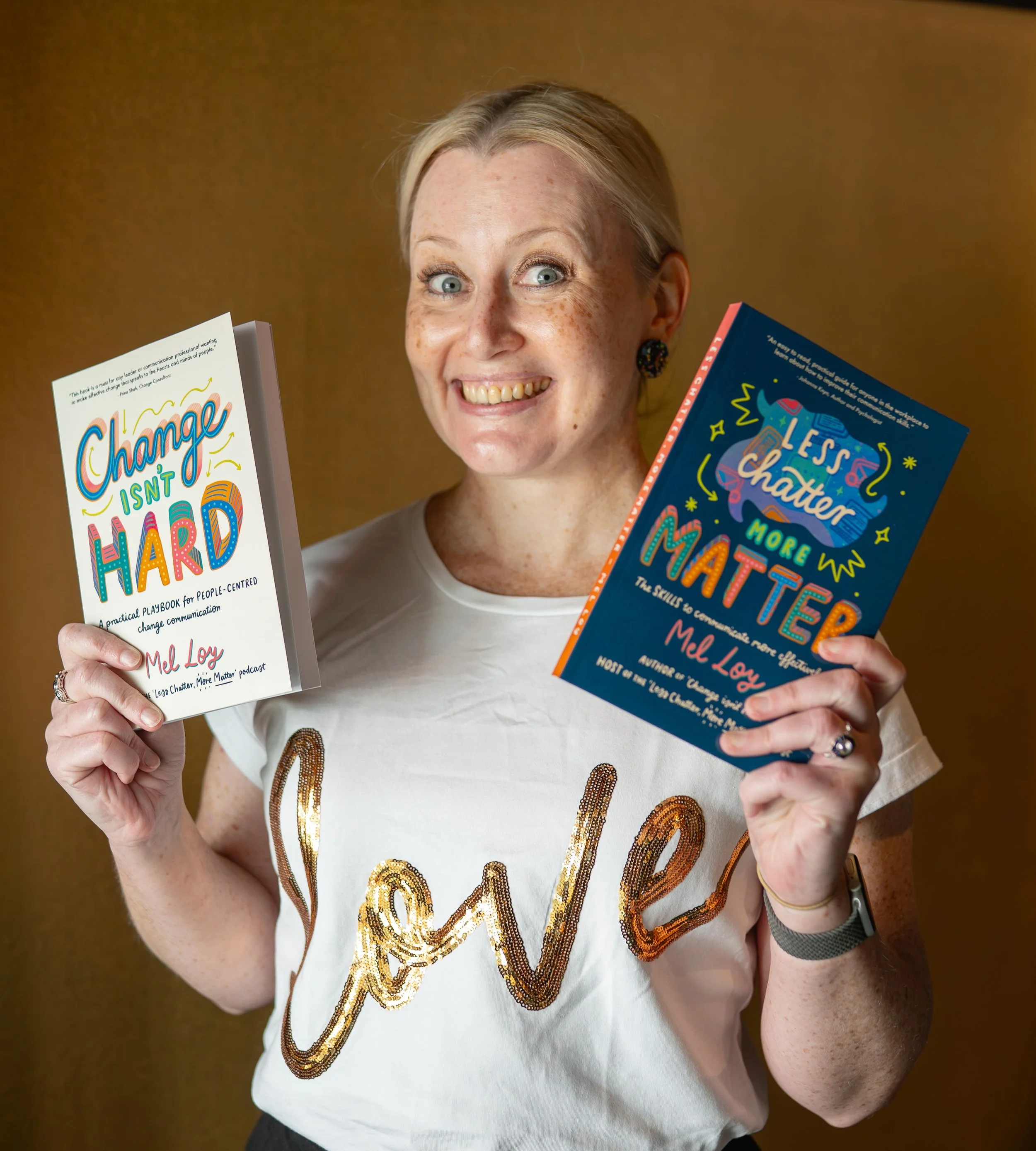 Photo of Mel smiling at the camera. She has short blonde hair pulled back, and wears a white t-shirt with the word 'love' in gold sequins on the front. She holds one of her books in each hand.
