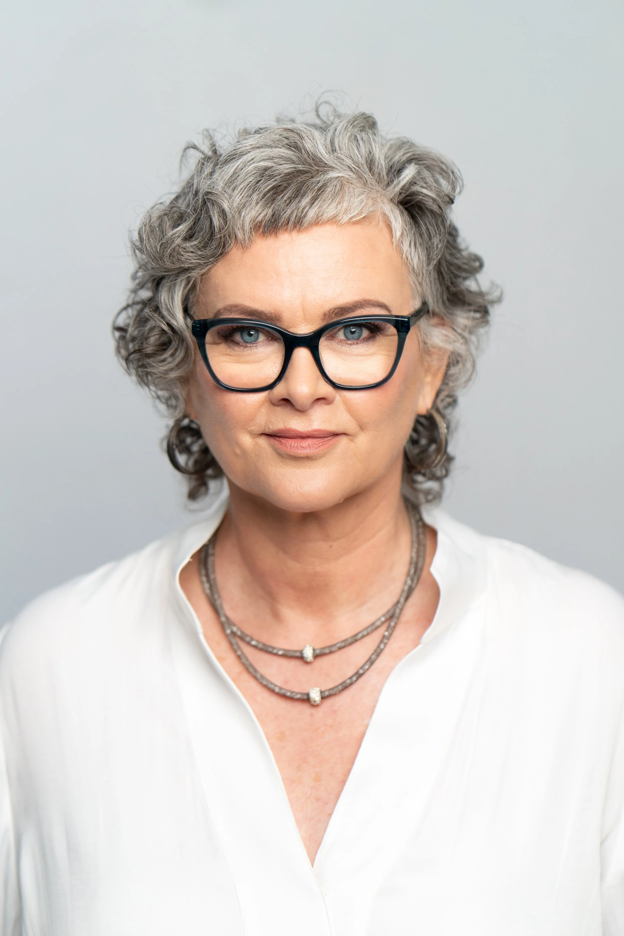 Photo of Jen smiling at the camera. She has short grey hair, wears large hoop earrings, a large silver necklace, and a white collared shirt.