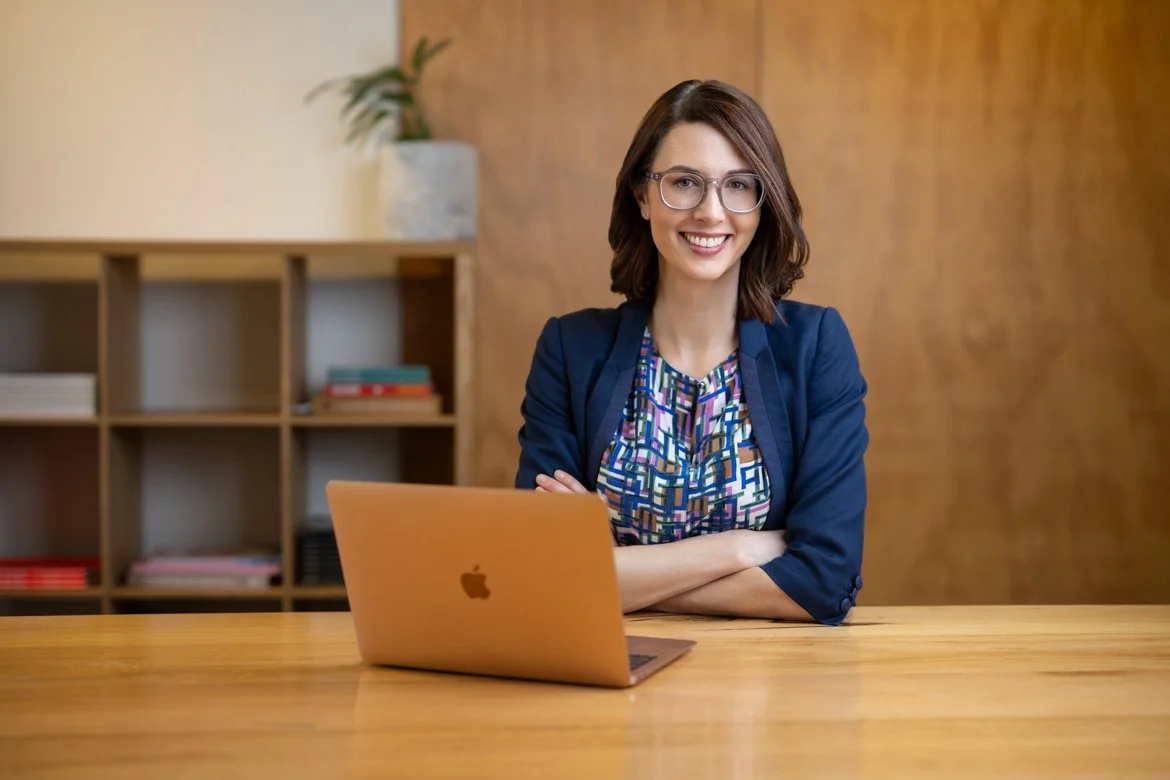 Photo of Julia, sitting a desk in front of a laptop, smiling at the camera. She has dark shoulder-length hair, glasses, and wears a blue blazer over a bright coloured shirt.