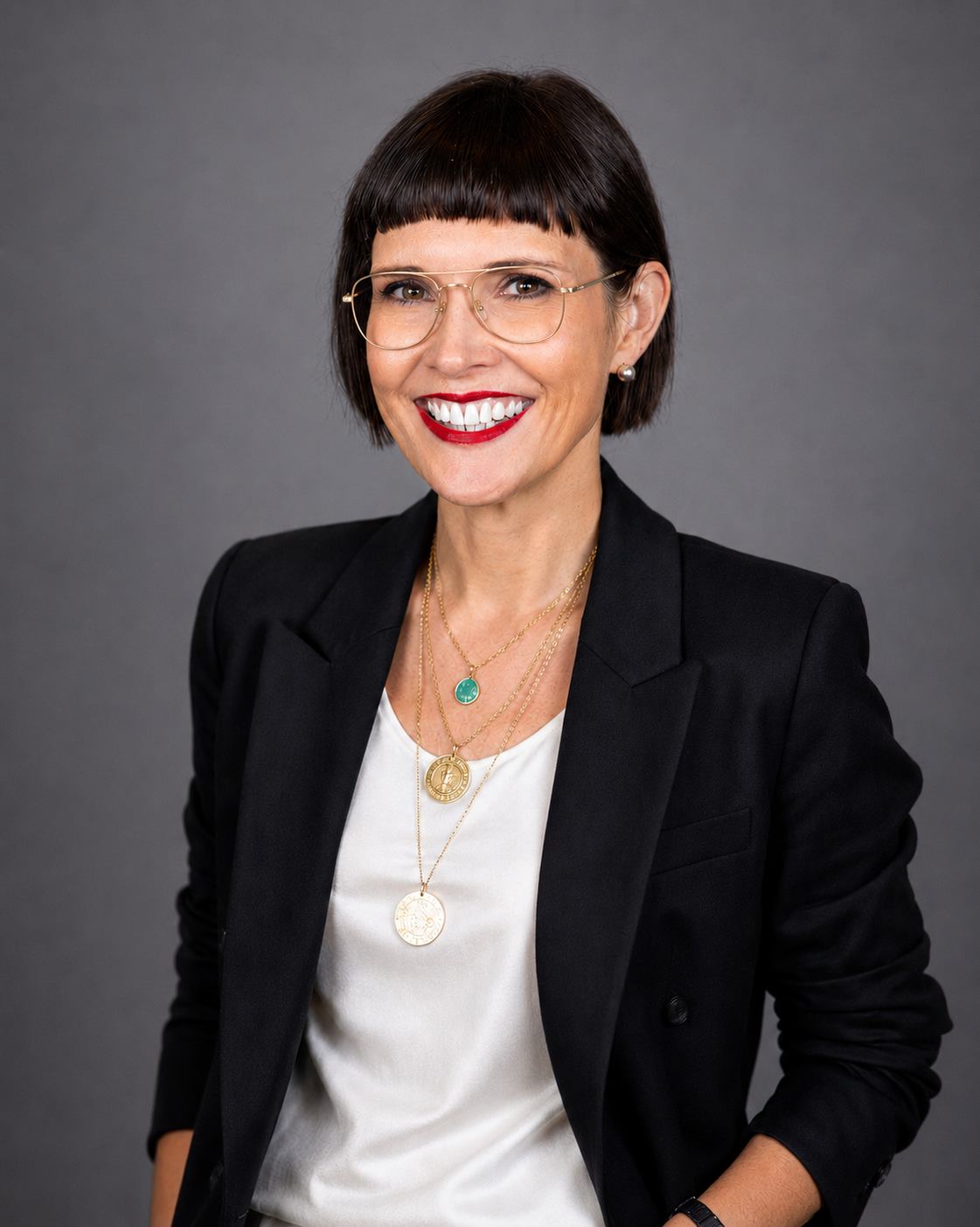 Photo of Angela smiling at the camera, she has short dark cropped hair, wears wire-rimmed glasses, a shite shirt with a black blazer.