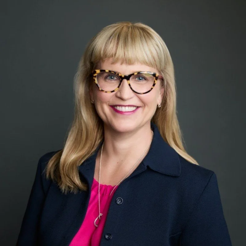 Photo of Katie smiling at the camera. She has long blonde hair and a fringe and wears tortoise shell-rimmed glasses, and a dark blazer over a bright pink shirt.