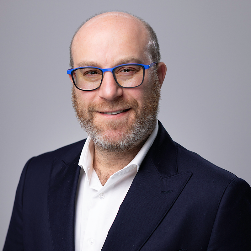 Photo of David smiling at the camera. He has a grey beard and moustache, wears glasses, and wears a dark-coloured jacket over a white collared shirt.