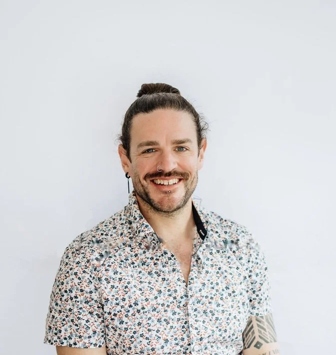 Photo of Shea smiling at the camera. He has his hair pulled back in a high bun, and has dark beard and moustache. He wears a paisley-print collared shirt.